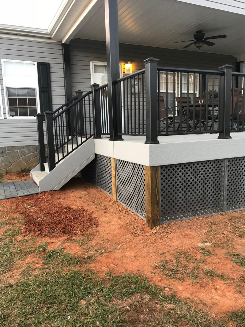 Gray and white porch with black railing, stairs, and lattice skirt. Red soil and grass in front.
