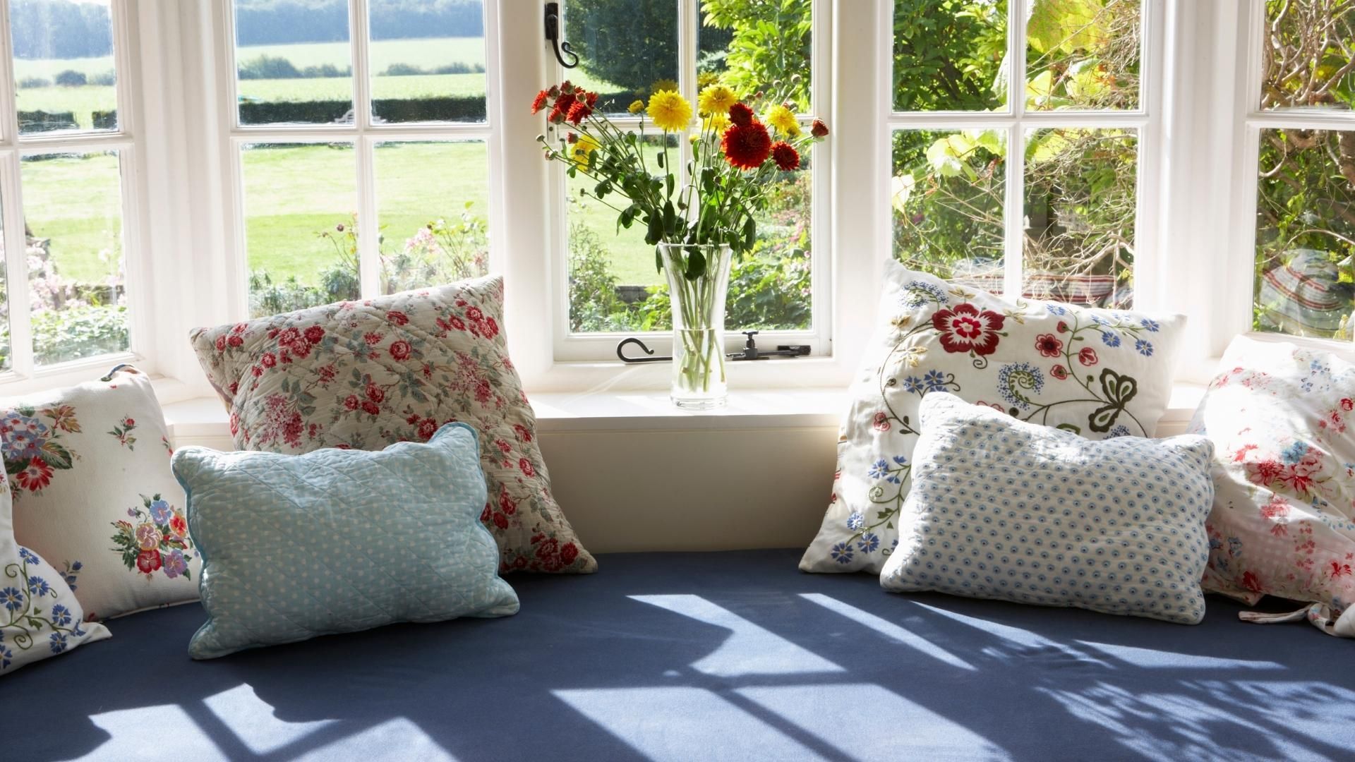 Window seat with floral pillows, blue cushion, and vase of flowers, overlooking a green landscape.