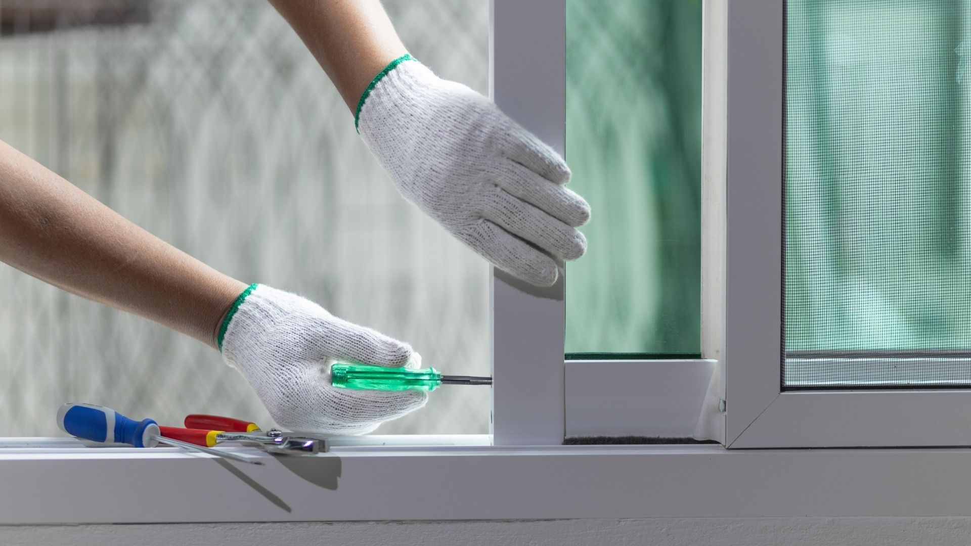 Hands in white gloves using a screwdriver to adjust a sliding glass door frame. Other tools are nearby.
