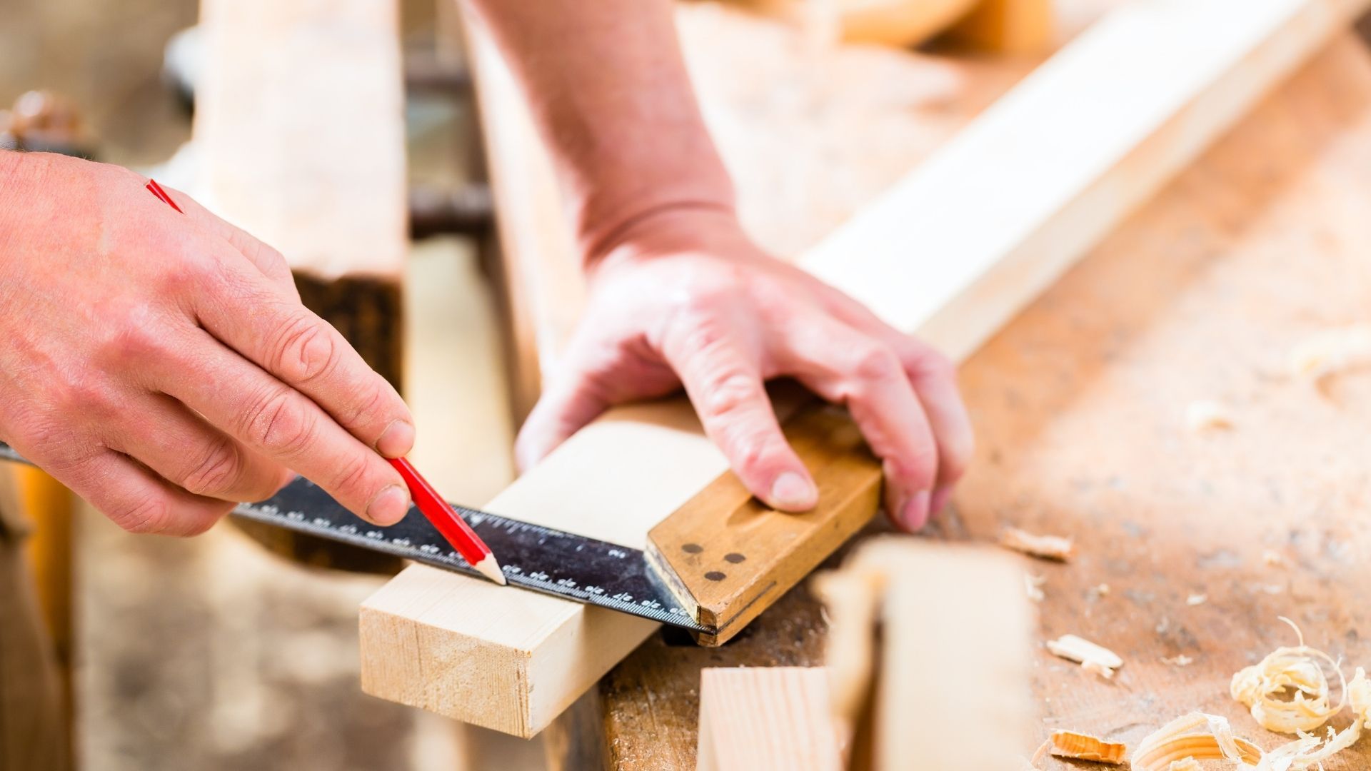 Person using a measuring square and pencil to mark a wooden plank. Workshop setting, sawdust visible.