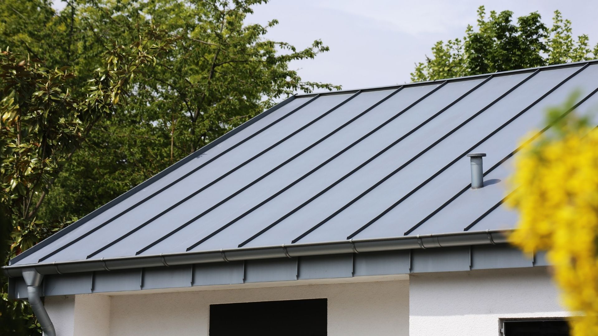 Gray metal roof on a white house with a gutter and a chimney against a backdrop of green trees.