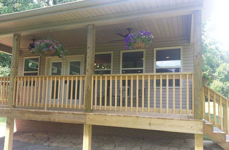 A porch with a wooden deck, railing, and roof. Hanging flower baskets and windows are visible.