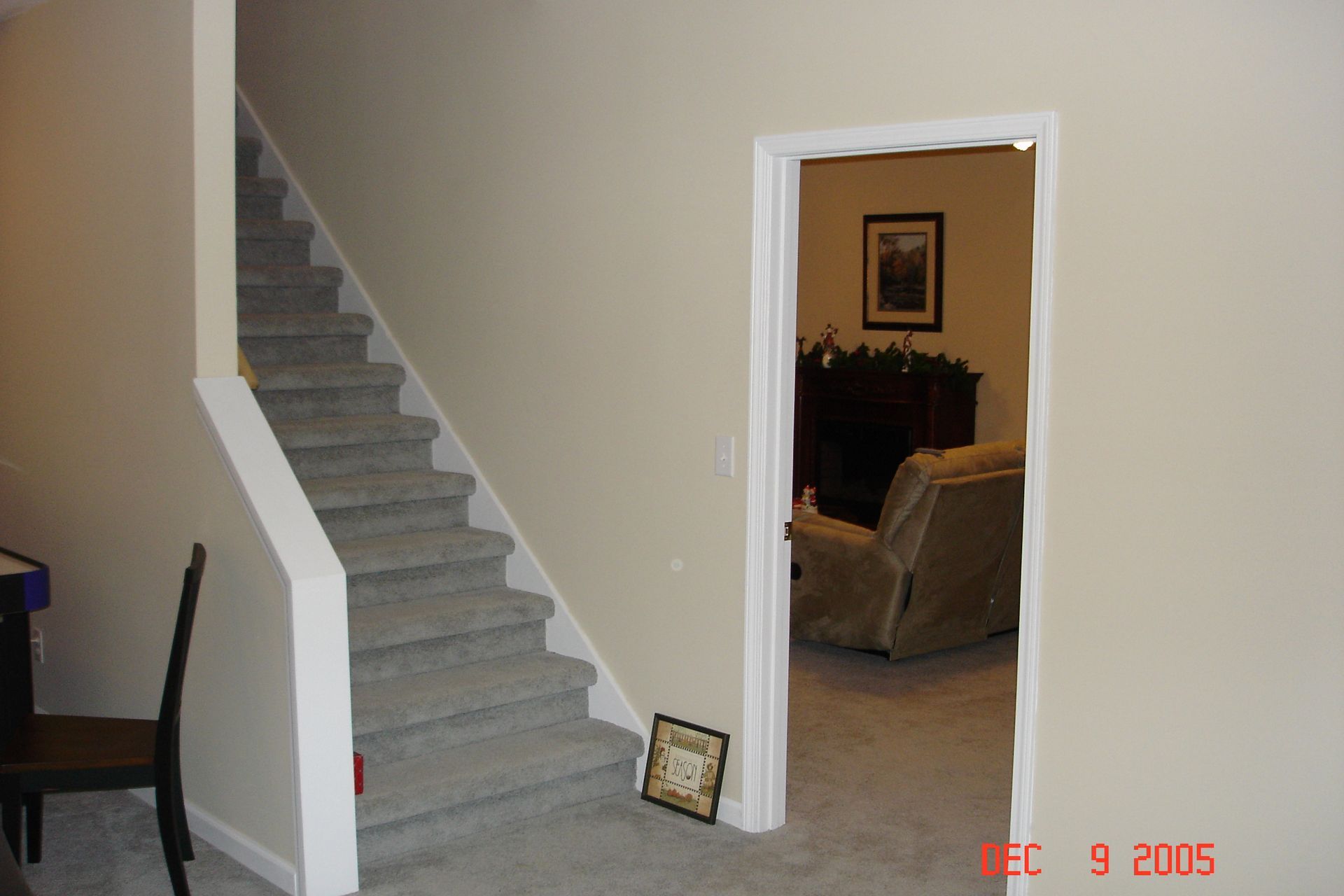 Staircase with gray carpet, leads up. Beige walls. Open doorway leads to living room with fireplace and recliner.