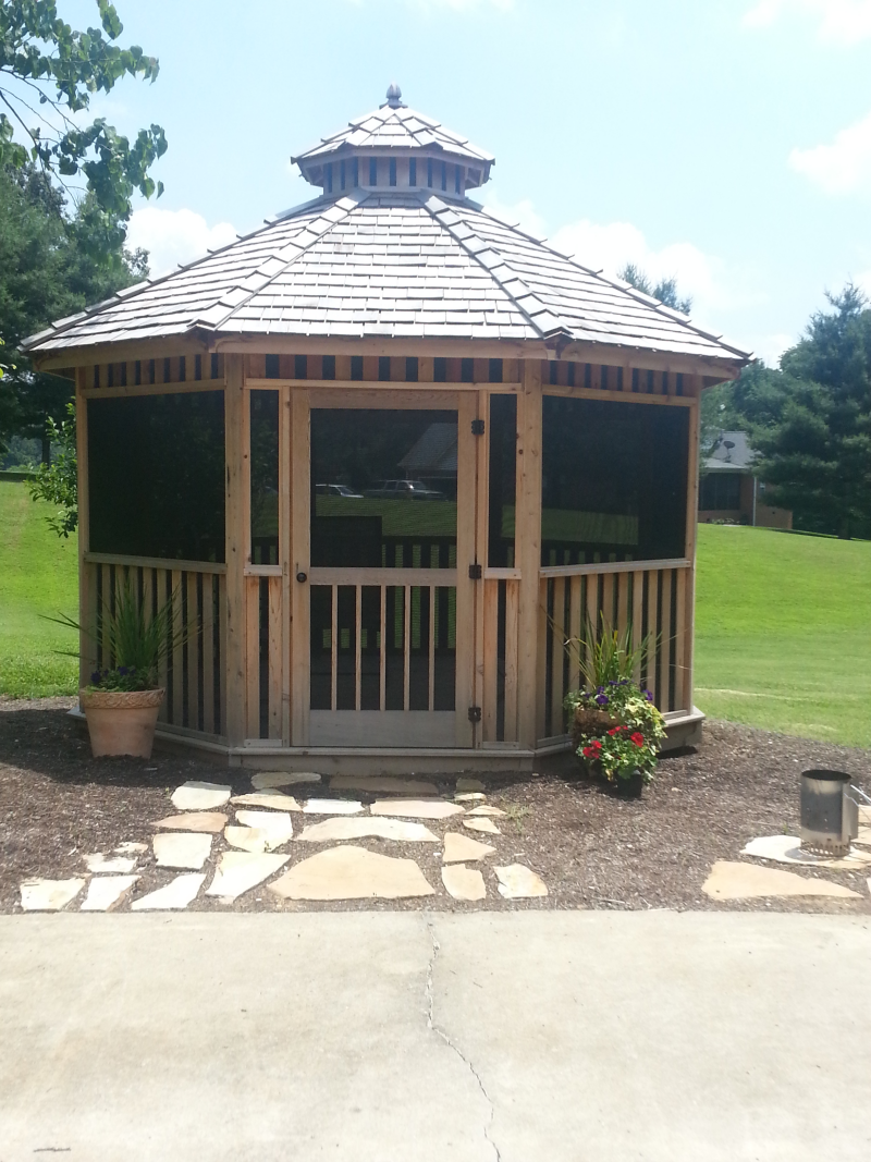 Wooden gazebo with screened walls, a wooden door, and a stone walkway.