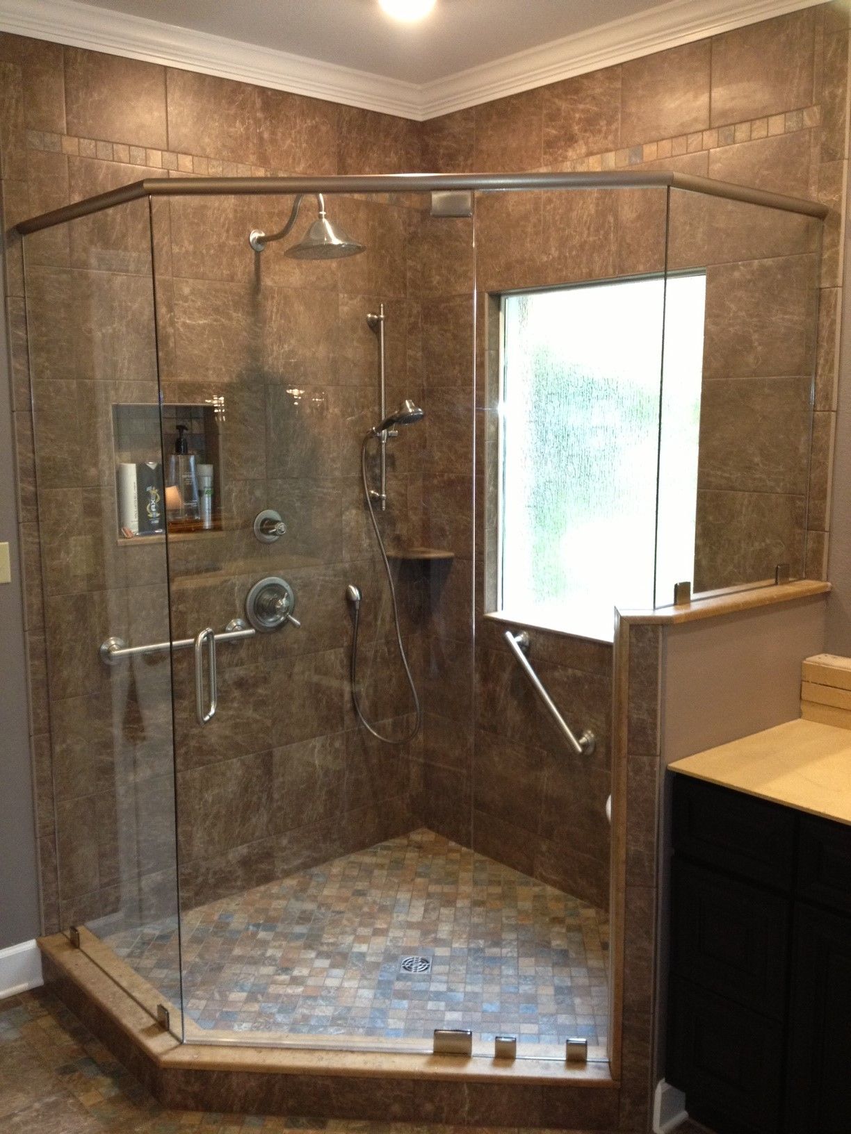Tiled shower with glass doors and grab bars, corner window. Earth-toned tiles, mixed floor tiles, chrome fixtures.