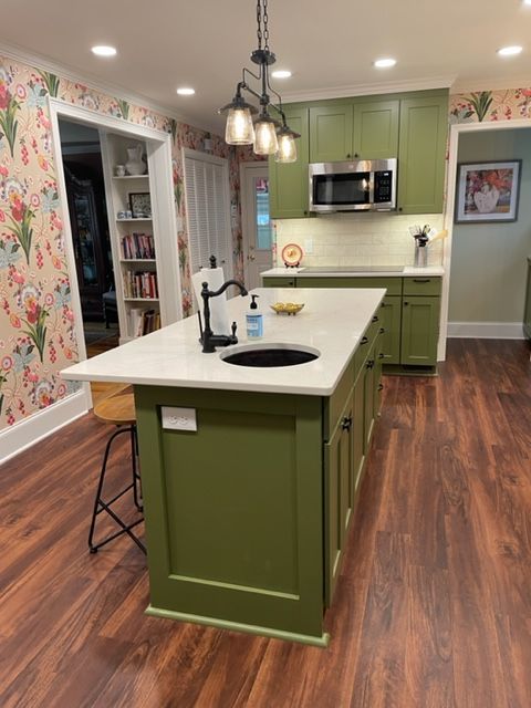 Green kitchen island with sink and countertop, surrounded by green cabinets, floral wallpaper, and wood flooring.