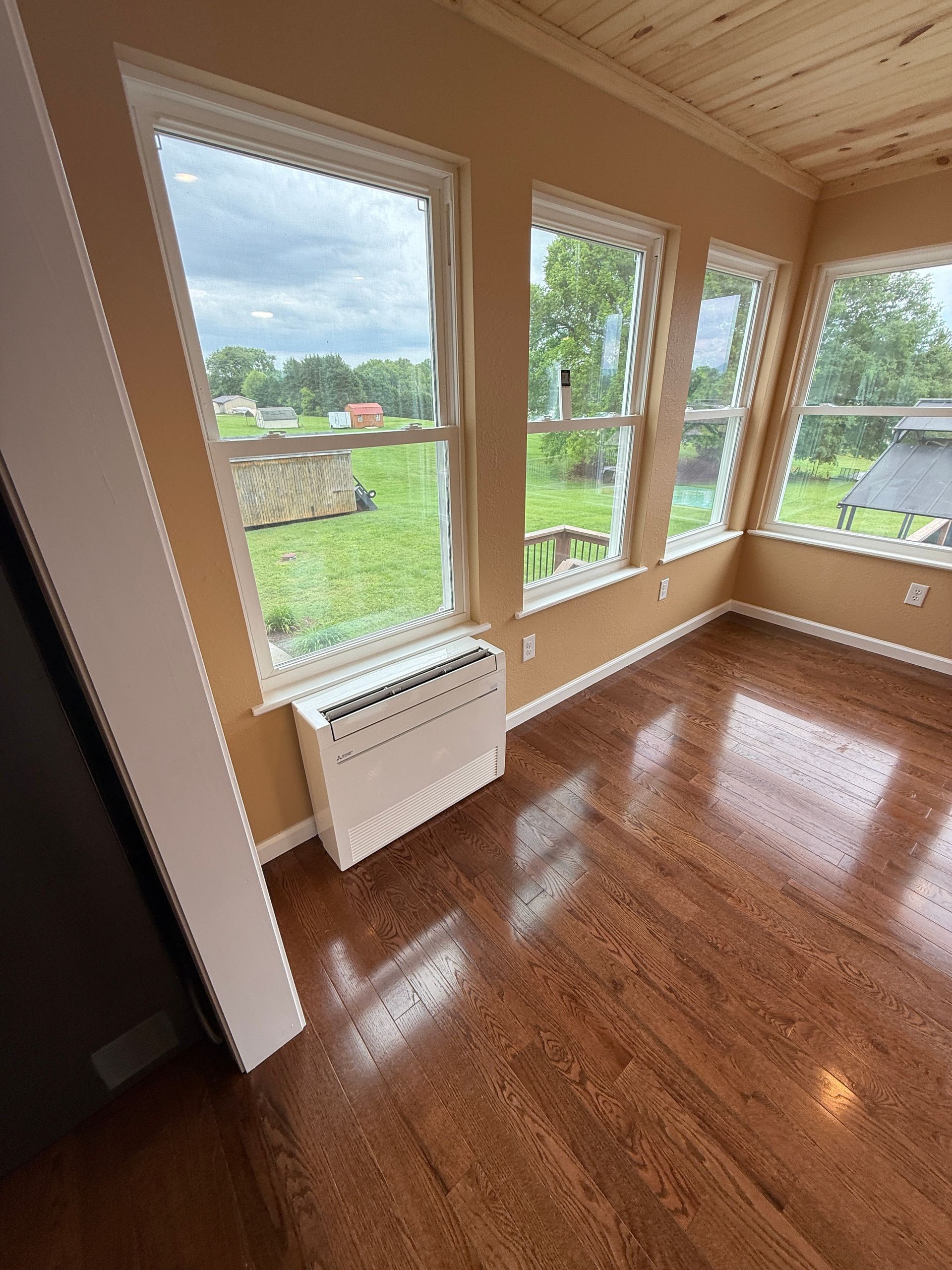 Interior view of a room with hardwood floors, windows, and an electric heater under a window, overlooking a green yard.