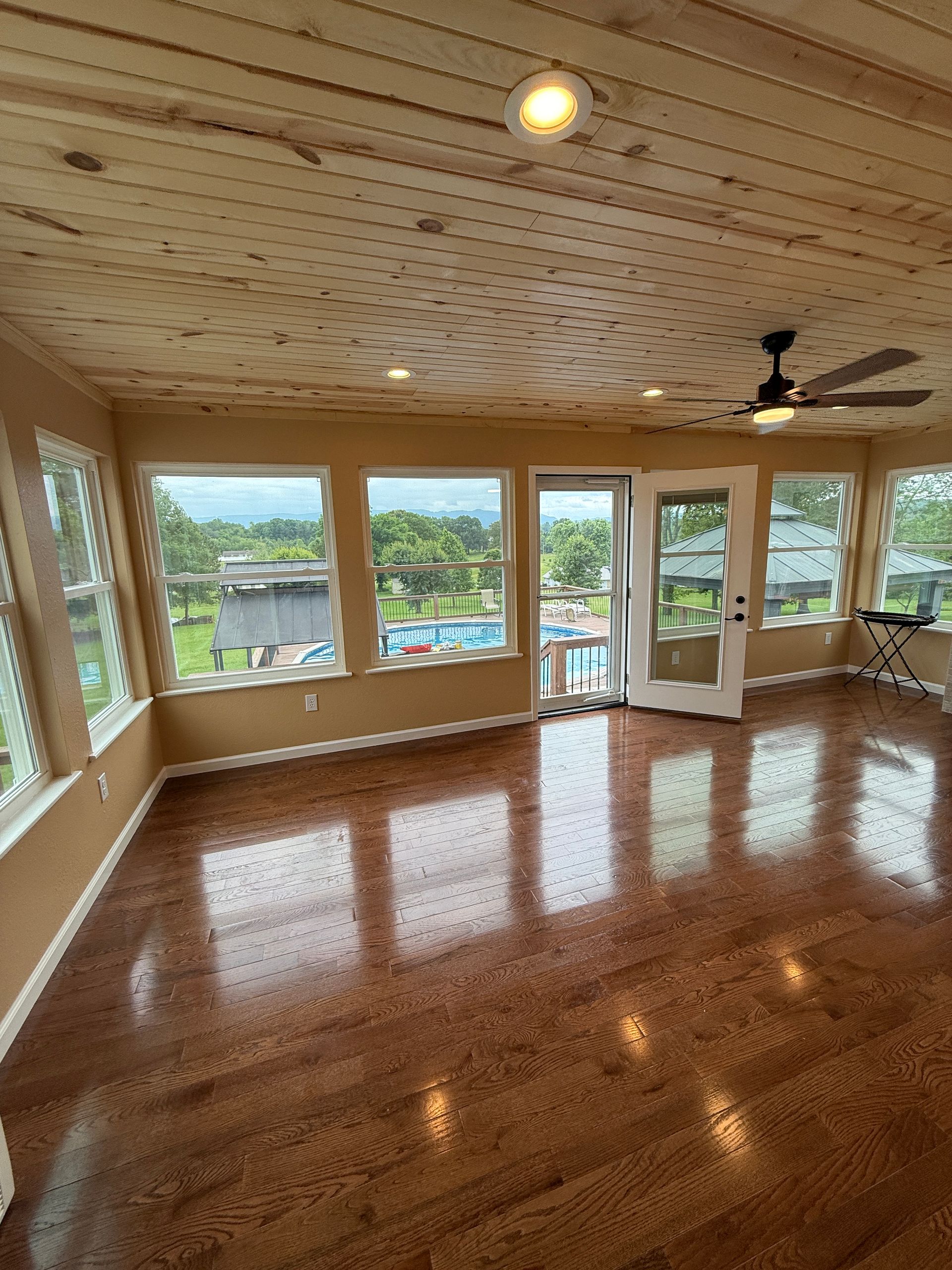 Sunroom with wood ceiling and floors, windows overlooking a pool and trees.