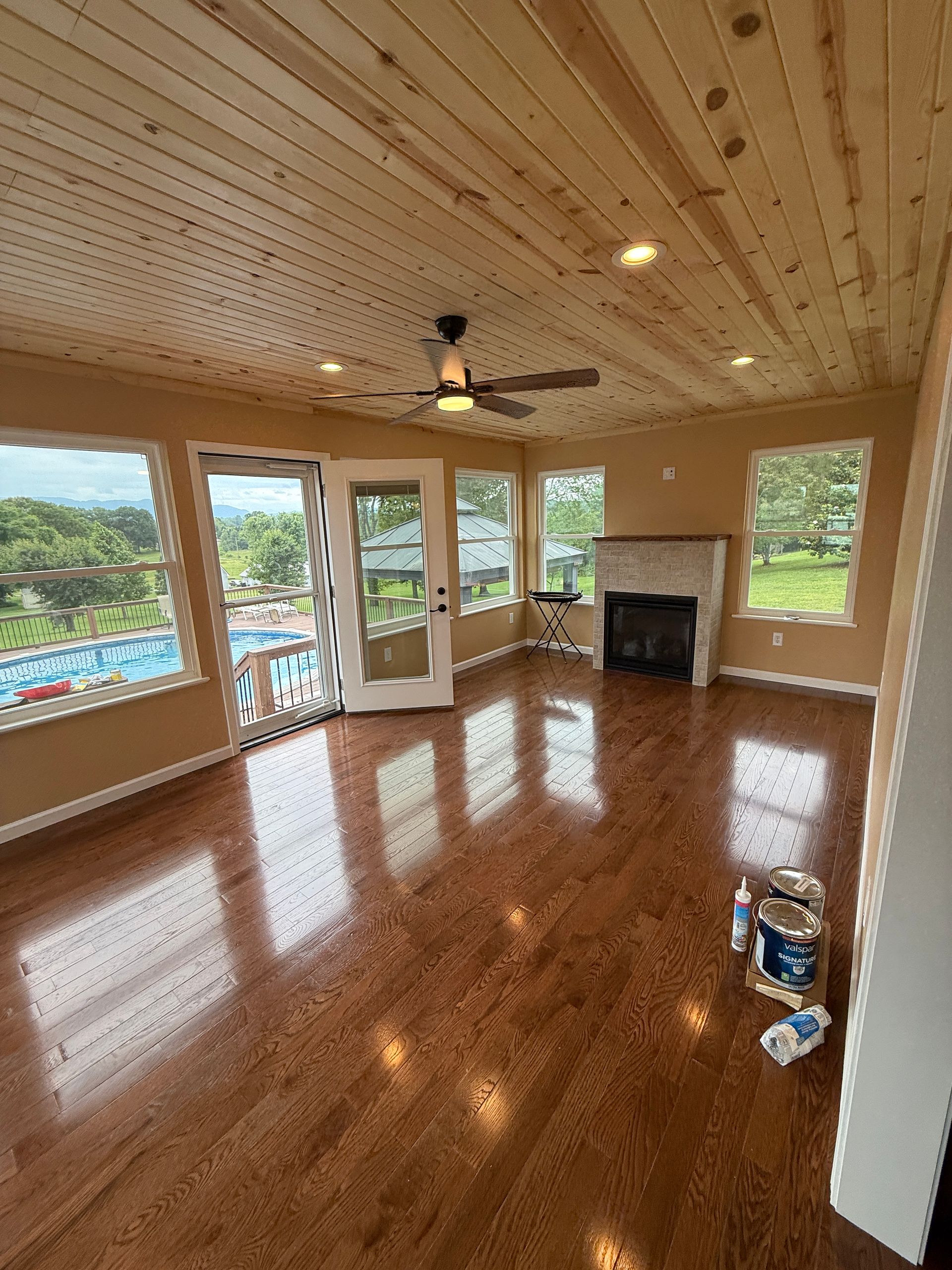 Spacious room with wood ceiling and floors, large windows, a fireplace, and a ceiling fan.