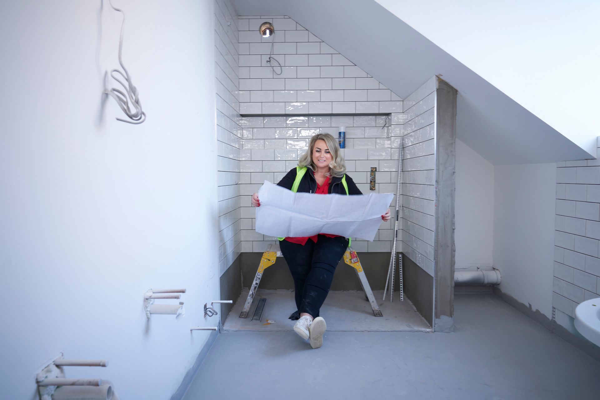 Woman in safety vest holds a blueprint in a bathroom under construction with tiled walls and a slanted ceiling.