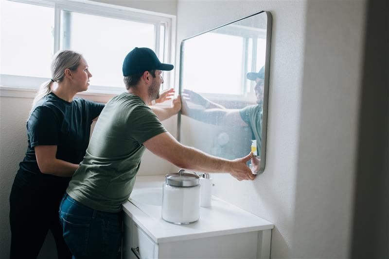 Couple hangs a mirror in a bathroom. The man holds it while the woman steadies it. White vanity and window in the background.