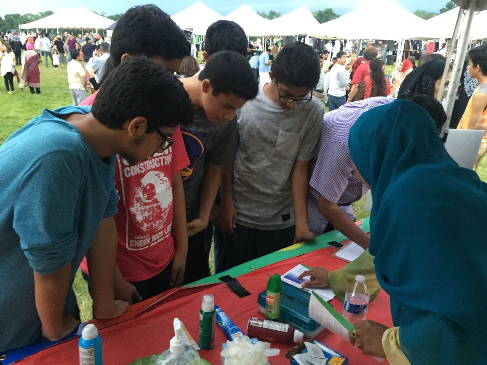 Group of people at a table, looking at items. Outdoors, cloudy day, tents in background.