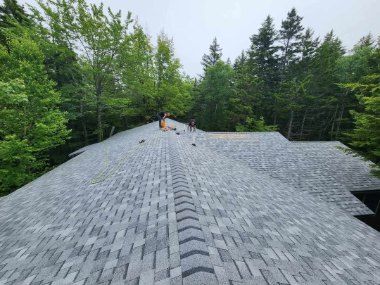 View of a newly shingled gray roof from the ridge looking down toward tree-lined woods.