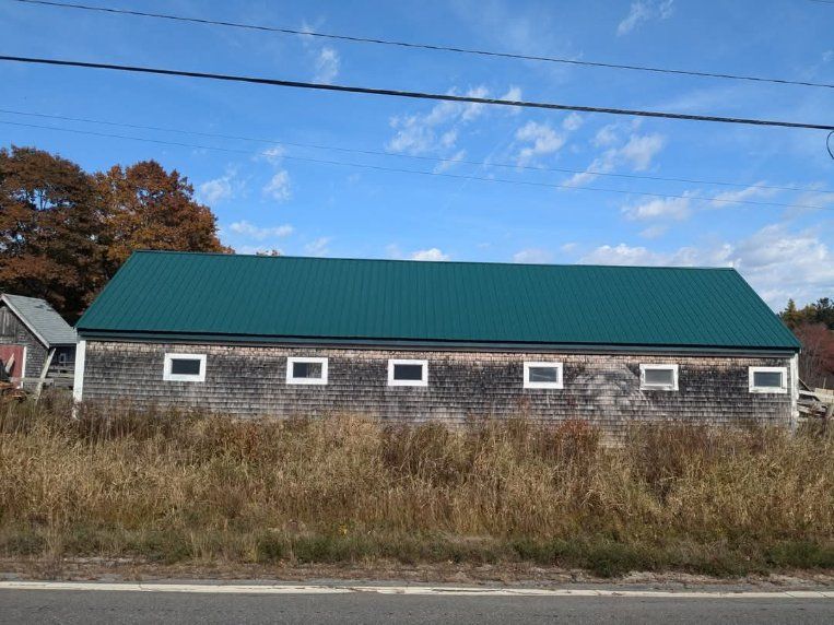A long, weathered wooden barn with a vibrant green metal roof and six small windows, set behind dry, brown autumn grasses.