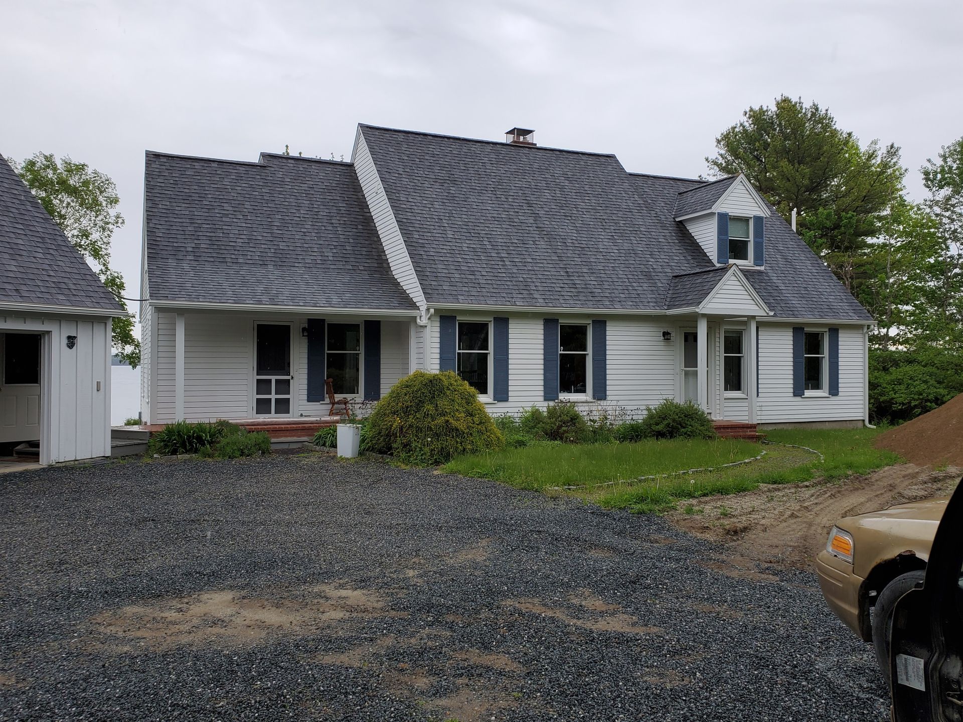 A white, single-story house with a grey, shingled roof and blue shutters, viewed from a gravel driveway.