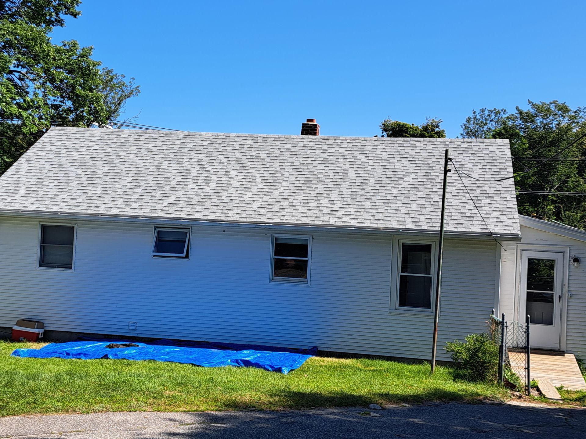 A white house with a gray shingled roof, three windows, a door on the right, and a blue tarp spread on the grass.
