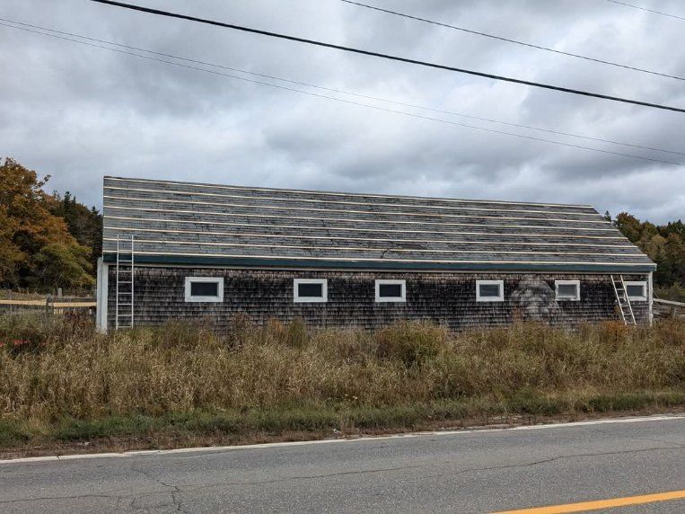A long, shingled barn with several windows sits behind tall grass next to an asphalt road under a cloudy sky.