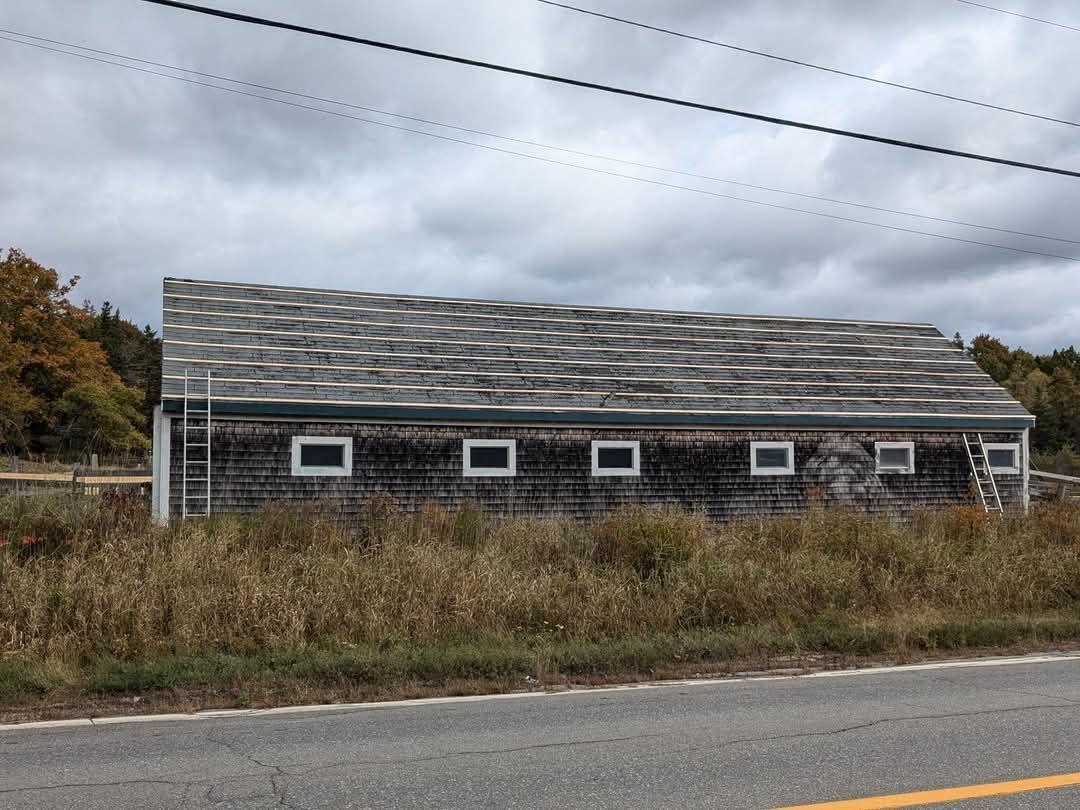 A weathered shed with a shingled roof and several small windows sits in a field beside a road under a cloudy sky.