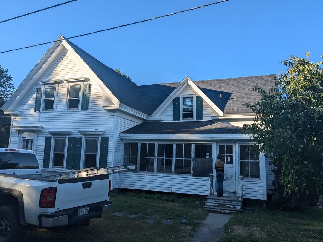 A white two-story house with a front porch, green shutters, and a parked white truck in front under a clear blue sky.