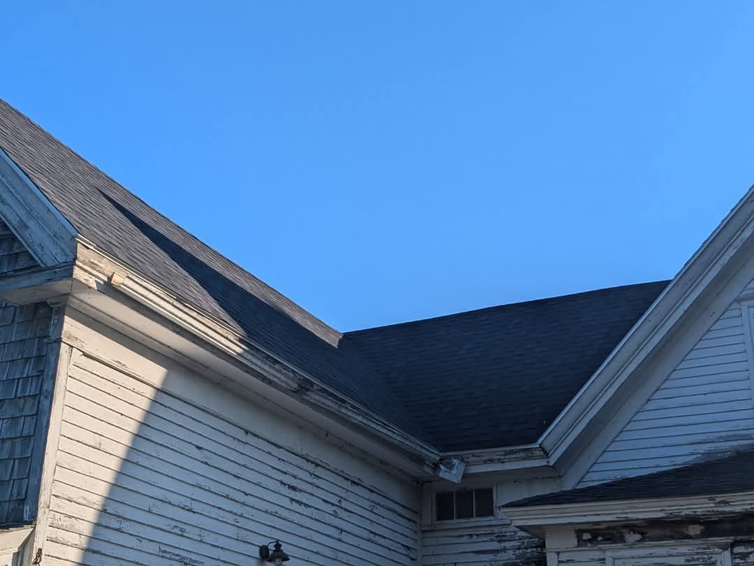 The roof and upper exterior walls of a white, weathered wooden house under a clear blue sky.