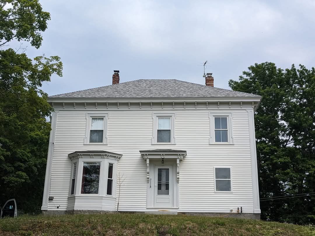 A two-story white house with a hip roof, bay window, and central entryway, surrounded by trees under a cloudy sky.