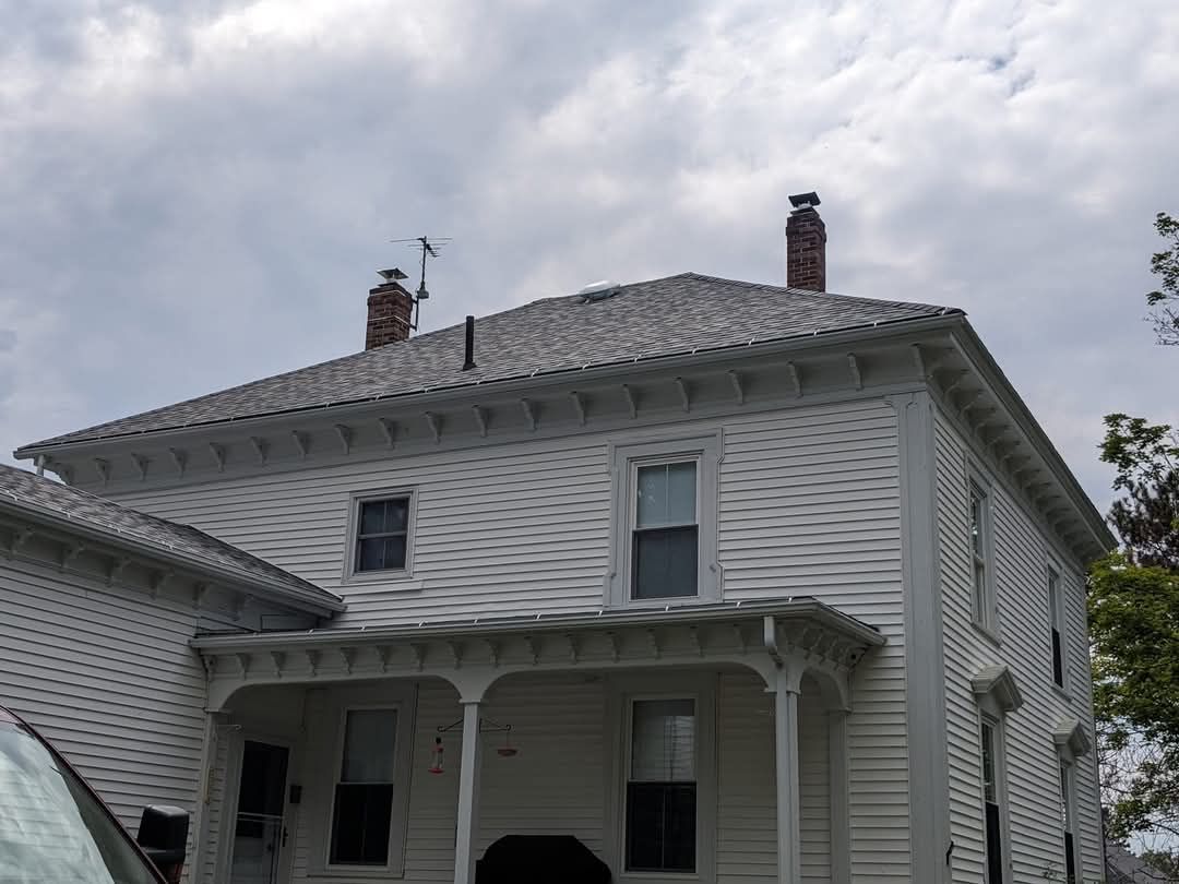 A two-story white house with clapboard siding, a dark roof with two chimneys, and a covered front porch under a cloudy sky.