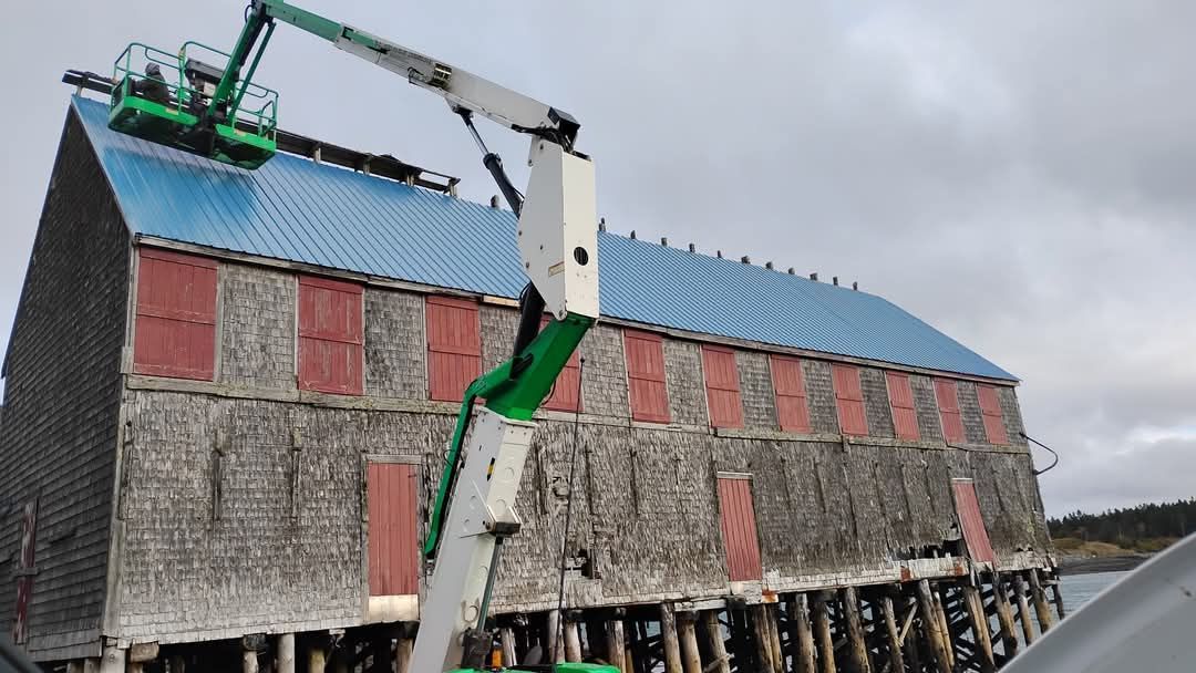 A green cherry picker lift works on the blue metal roof of an old, weathered wooden building on pilings near the water.