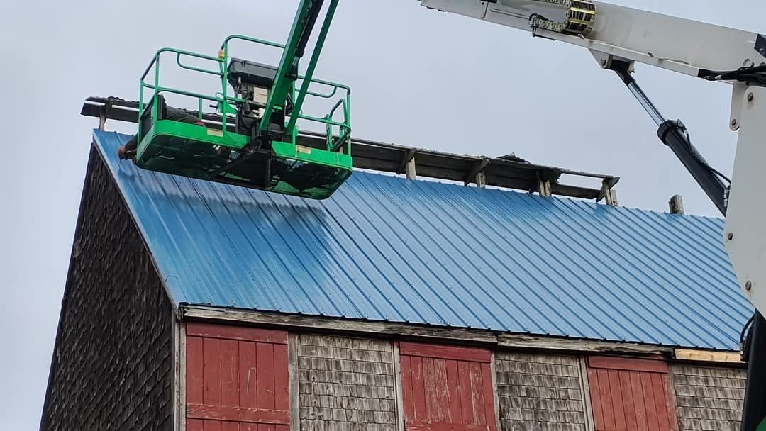 A green aerial lift basket hangs over a blue metal roof on an old building with weathered wood siding.