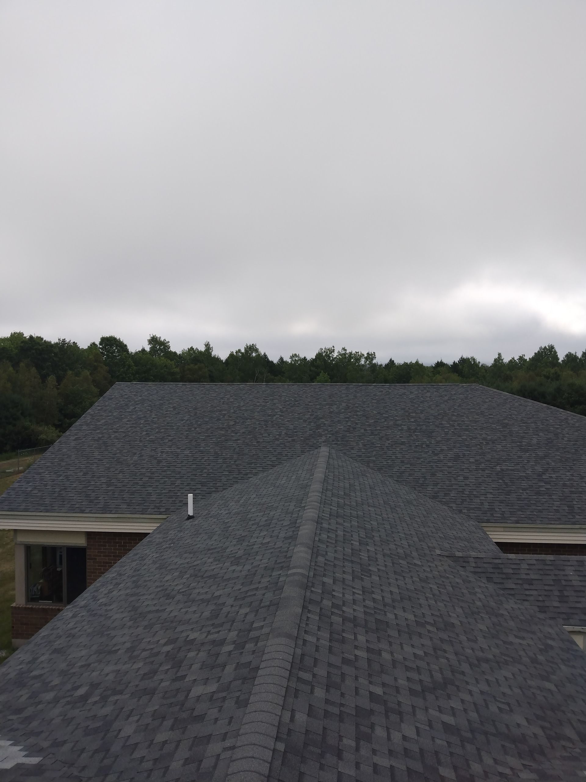 A high-angle view of a grey shingled roof under a cloudy sky, with trees in the background and a chimney pipe.