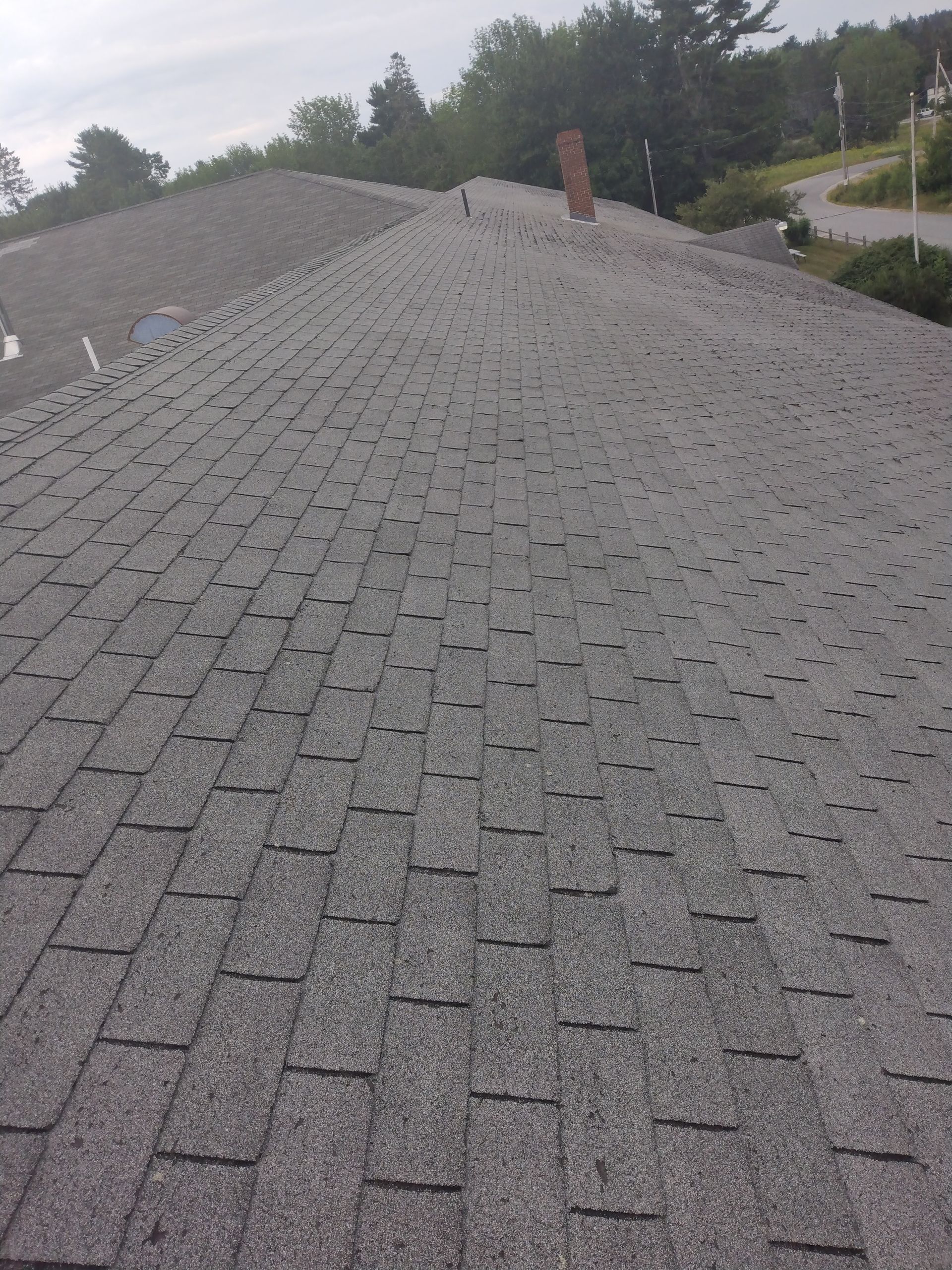 A top-down view of a grey asphalt shingle roof, showing a chimney near the ridge and a suburban setting in the background.