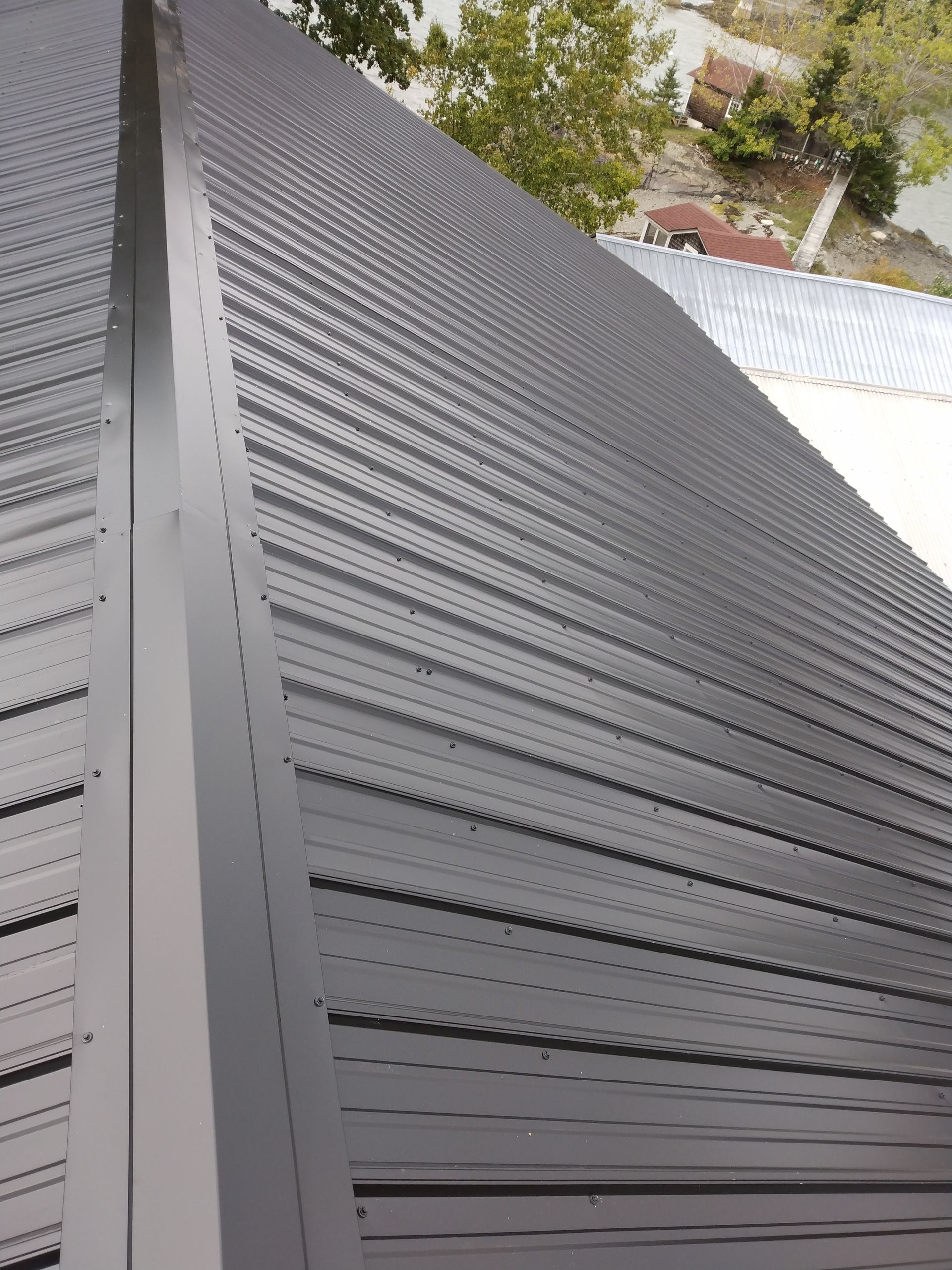 A high-angle view of a dark grey metal roof with a central ridge cap, showing the textured panels and outdoor scenery.
