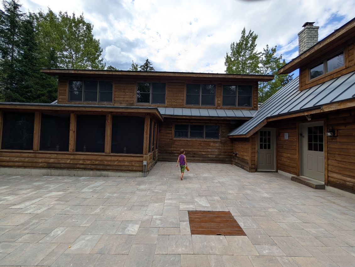 A child walks across a stone patio toward a large, rustic log cabin with a metal roof and a screened-in porch.