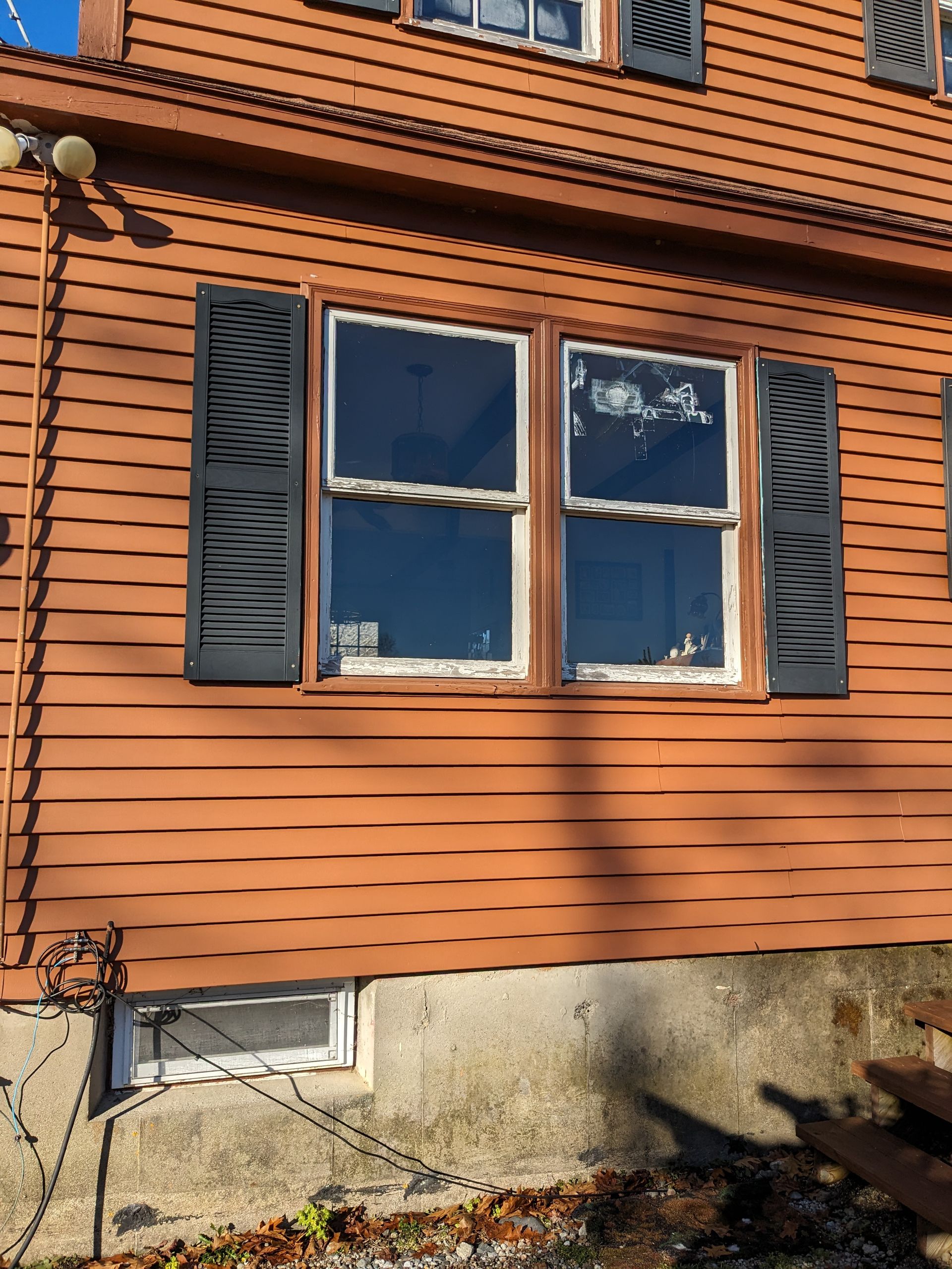A tan house exterior with black shutters flanking a double window above a concrete foundation and a small basement window.