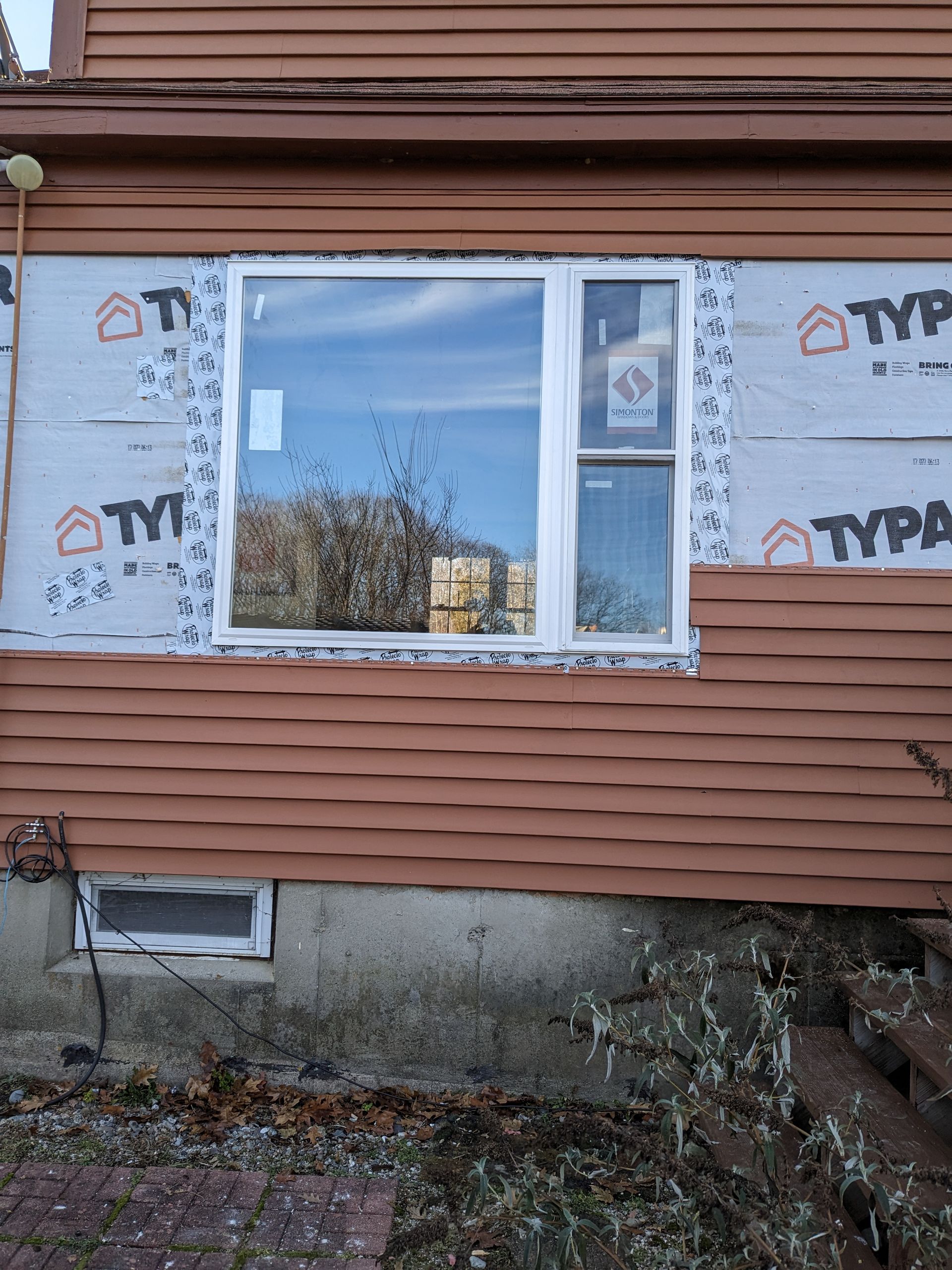 A white-framed window installed in a brown-sided house wall with exposed silver weather-resistant building wrap.