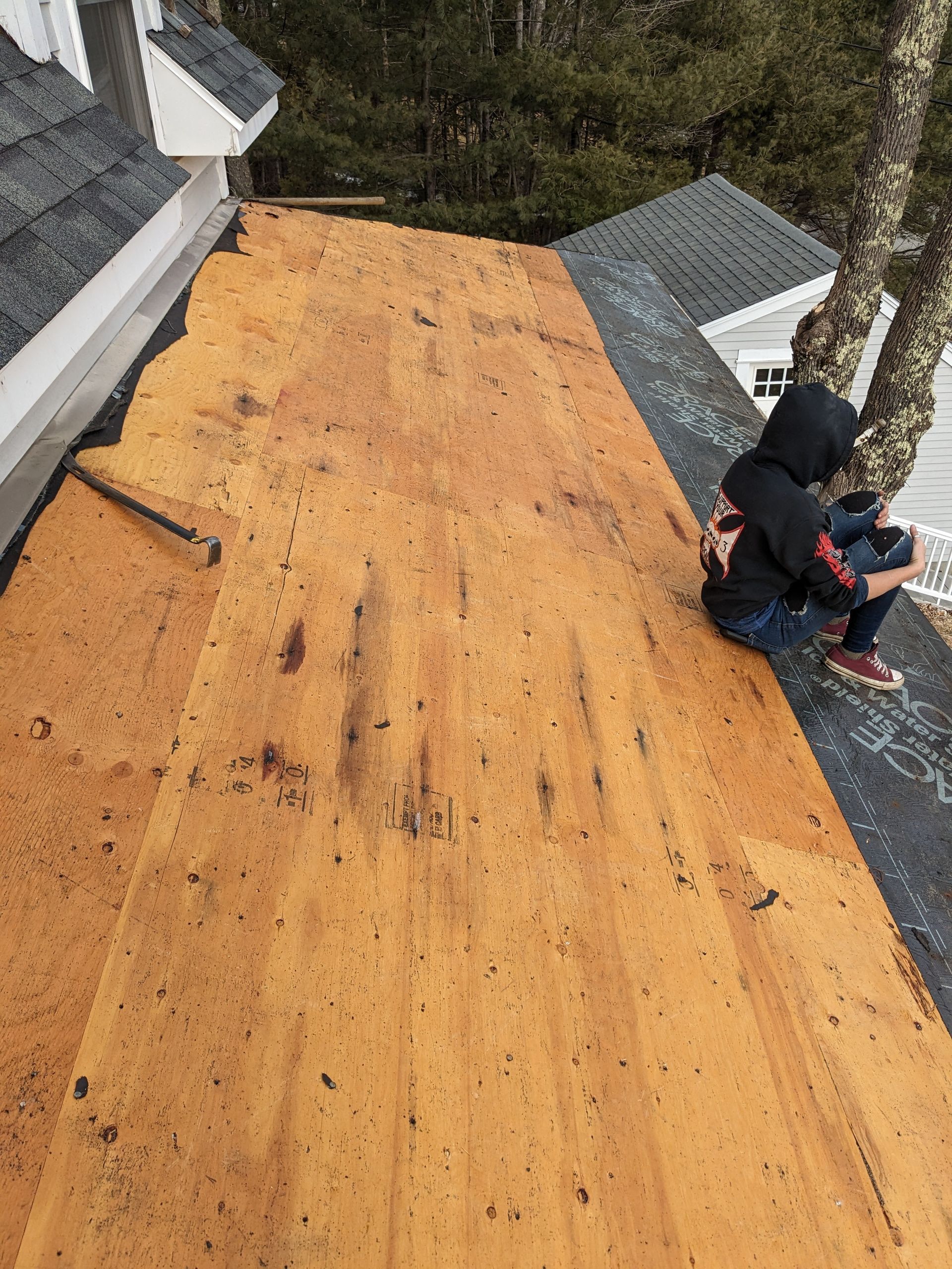 A person in a black hooded sweatshirt sits on a wooden roof deck under repair, with shingles visible nearby.
