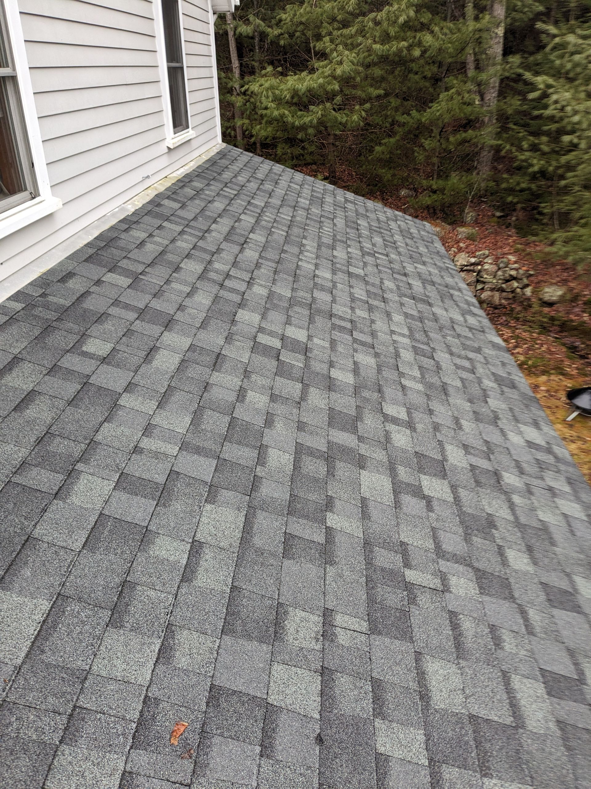 A high-angle view of a grey architectural shingle roof extending from the side of a house toward a wooded area.