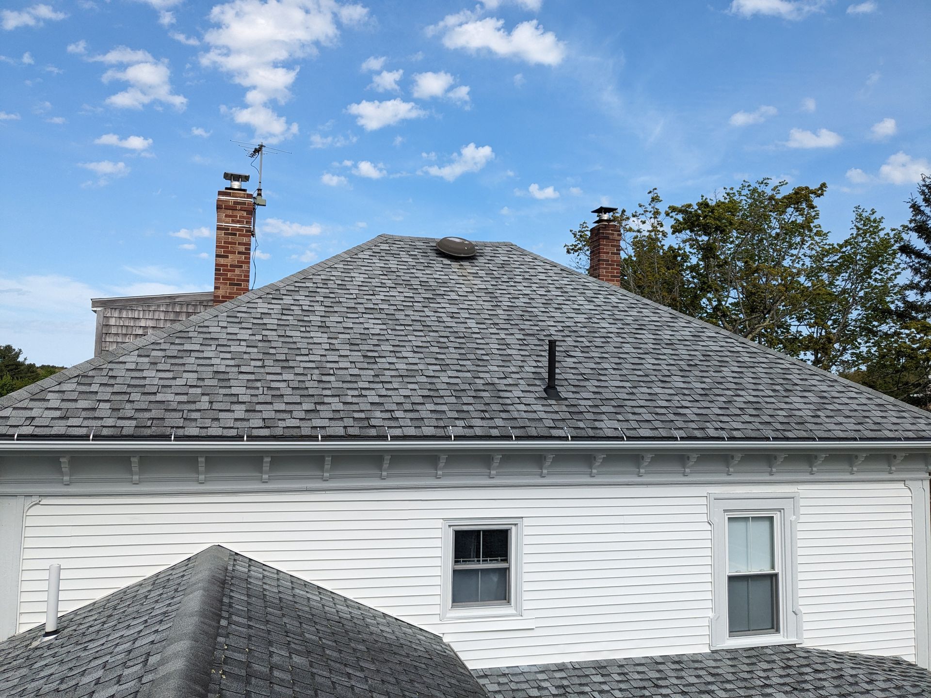 A exterior view of a white, shingled two-story house with two brick chimneys, asphalt roof shingles, and blue sky.