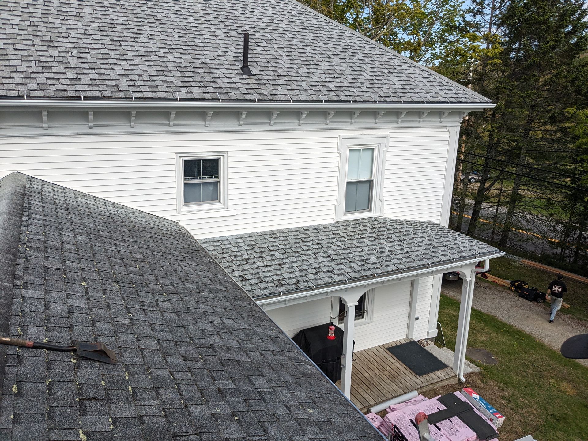 An elevated view of a house with a newly shingled grey roof, white siding, and a wooden porch with construction materials.