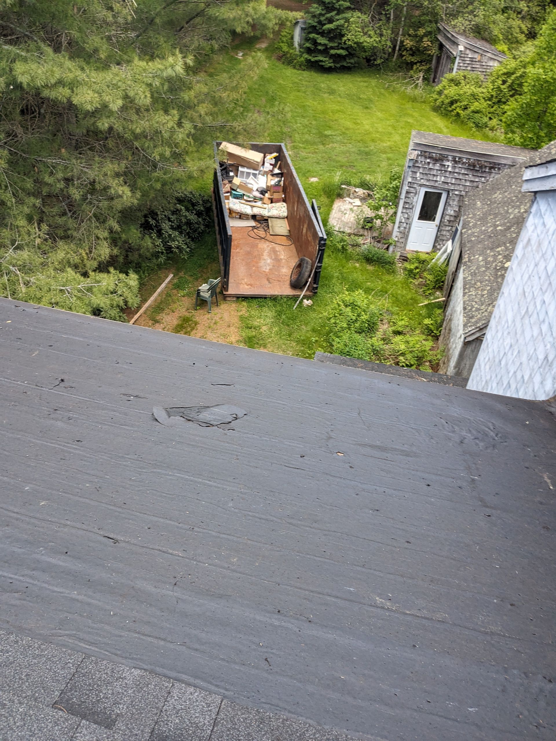 High-angle view of a residential roof slope leading down toward a dumpster filled with debris in a backyard.