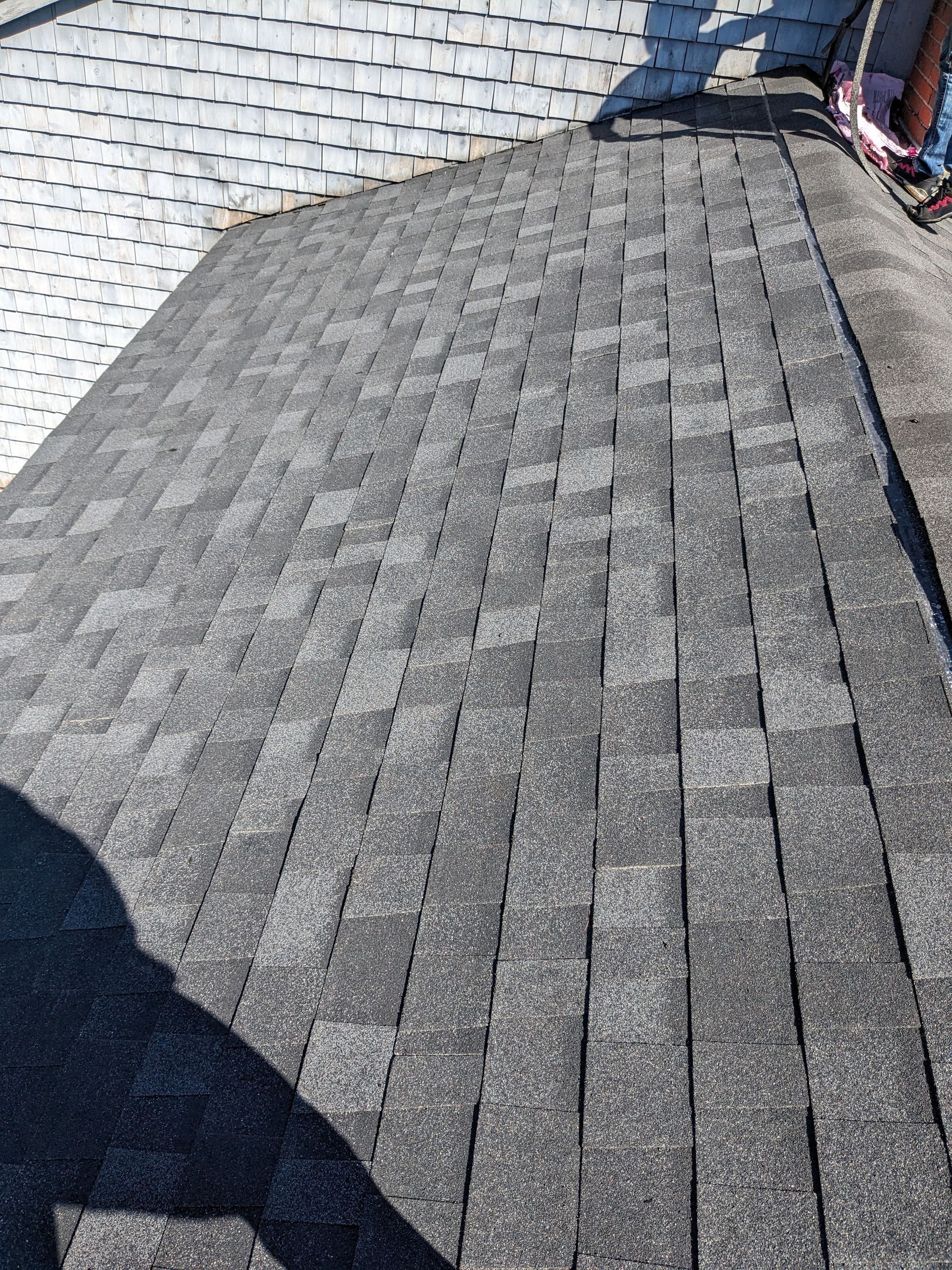 A view looking down a pitched roof covered in grey architectural asphalt shingles under bright sunlight.