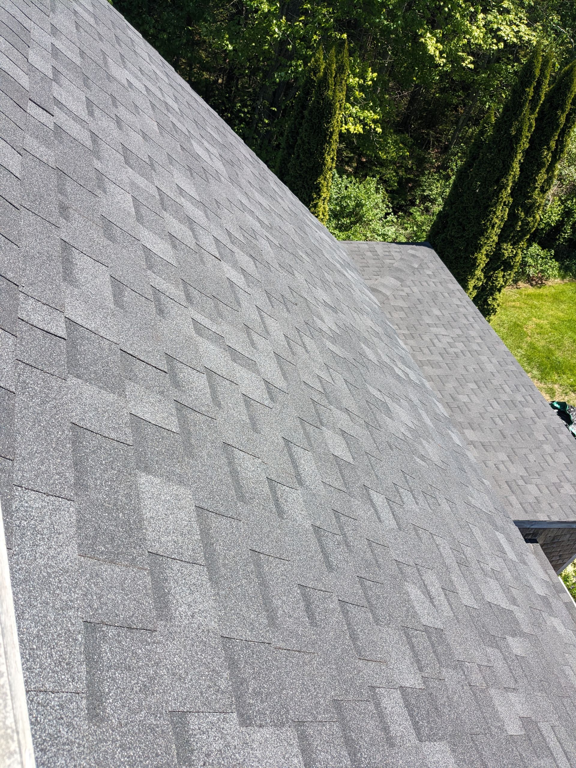 A high-angle view of a gray asphalt shingle roof, with a glimpse of green trees and grass in the background.