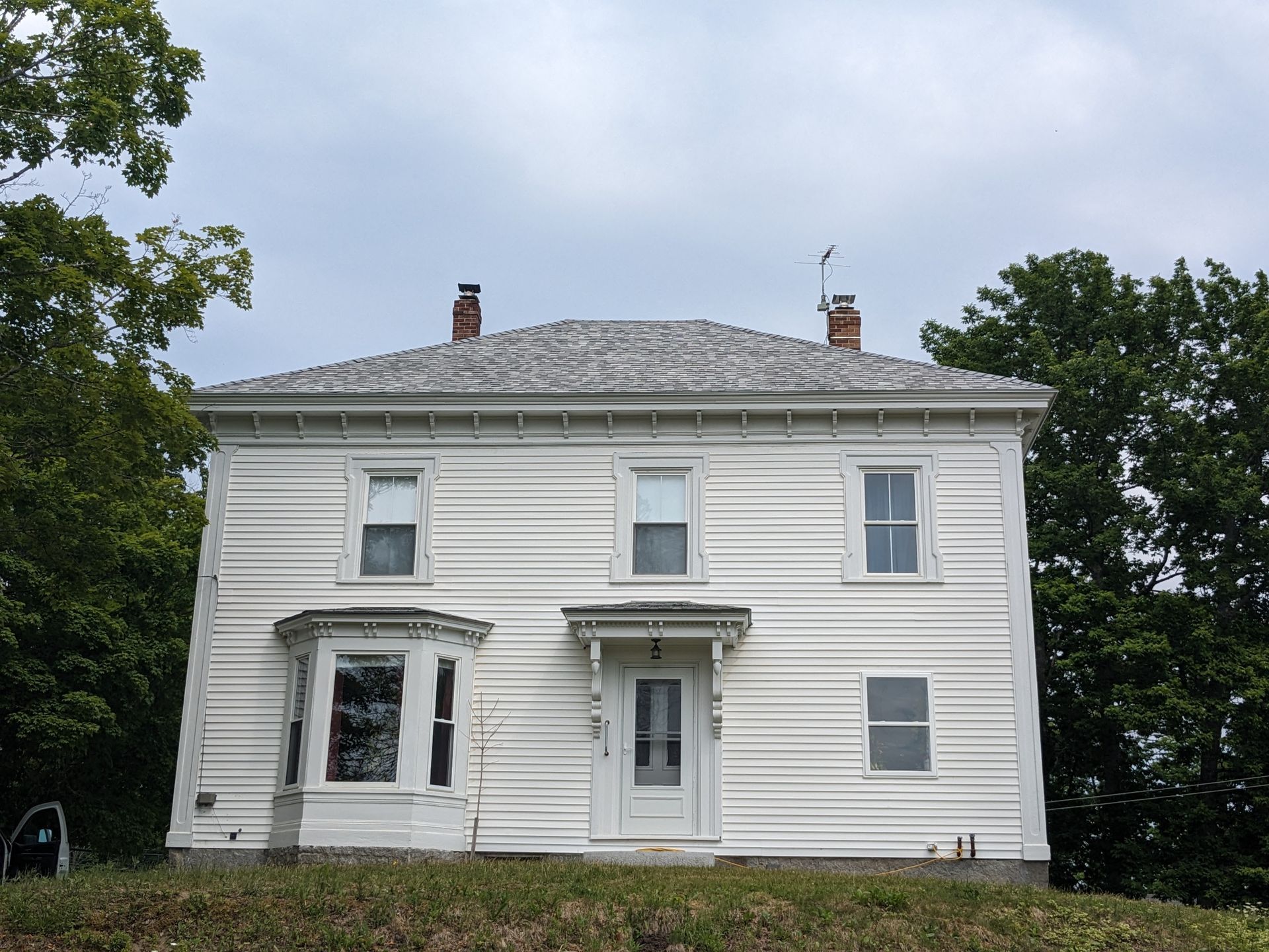 A two-story, white wood-sided house with a central front door, a bay window on the left, and trees on both sides.