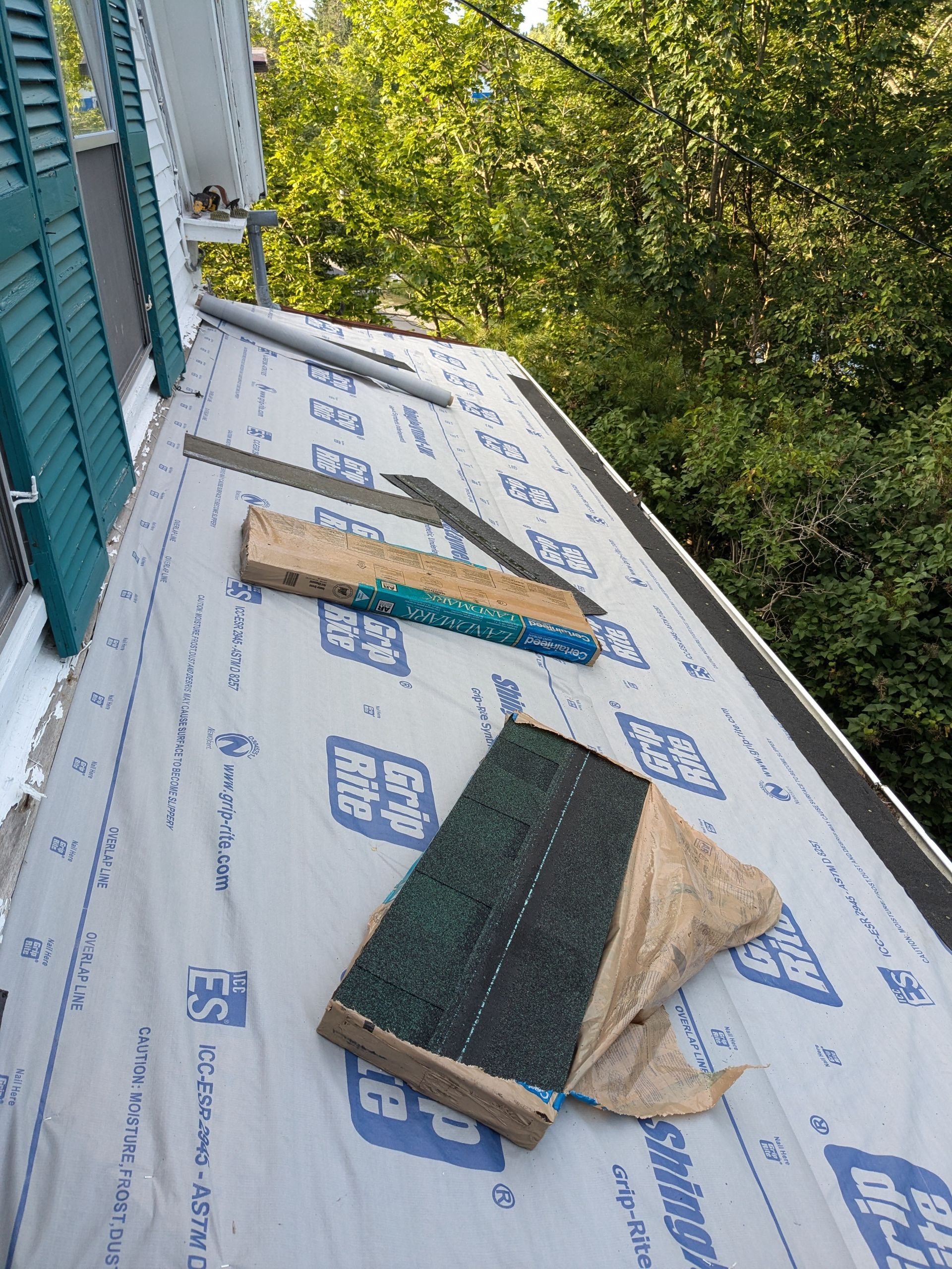 A low-angle view of a residential roof under repair, covered in grey underlayment with scattered tools and a roofing shingle.
