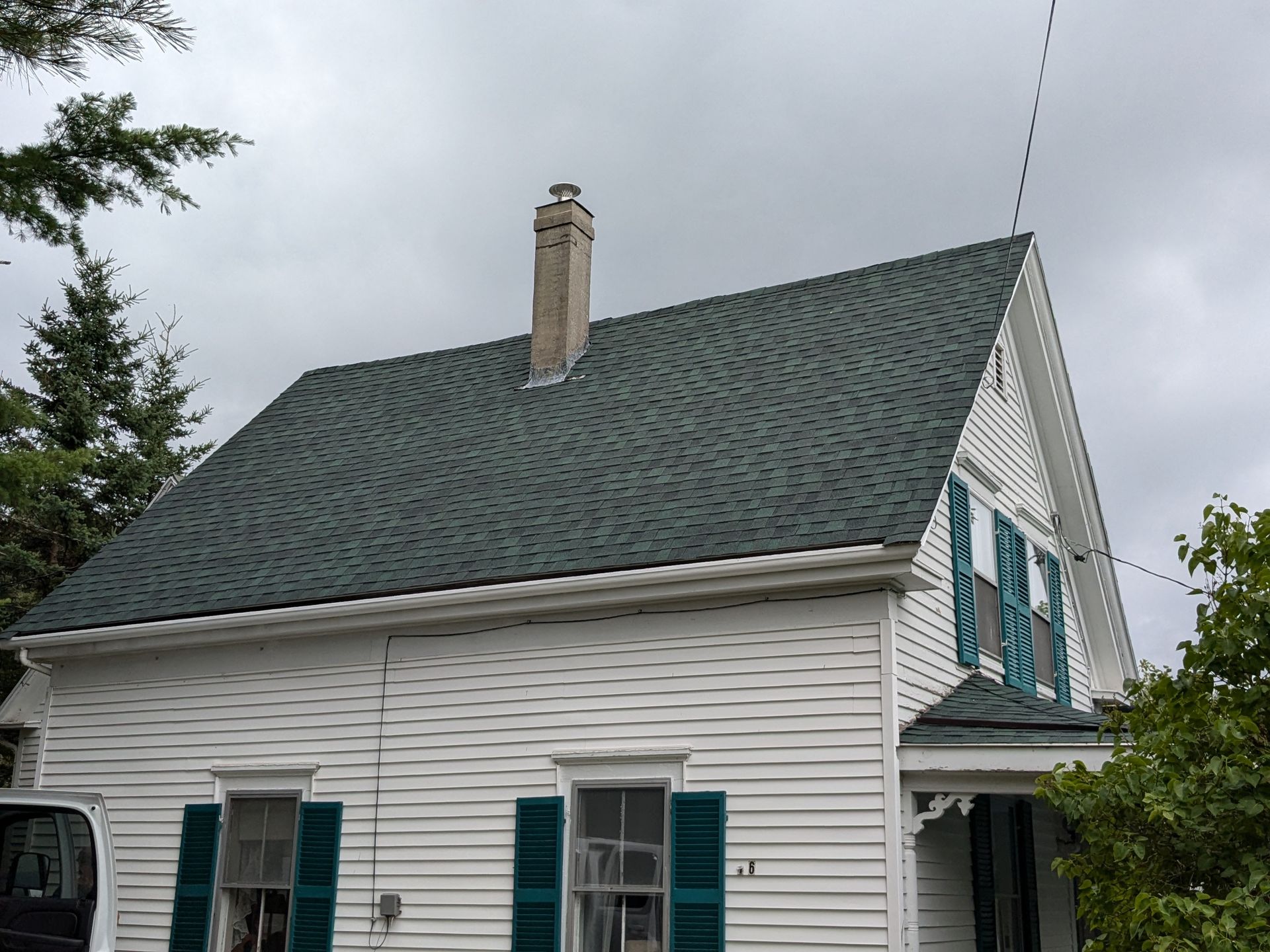 A white two-story house with green shutters, a gray shingled roof, and a stone chimney under a cloudy sky.