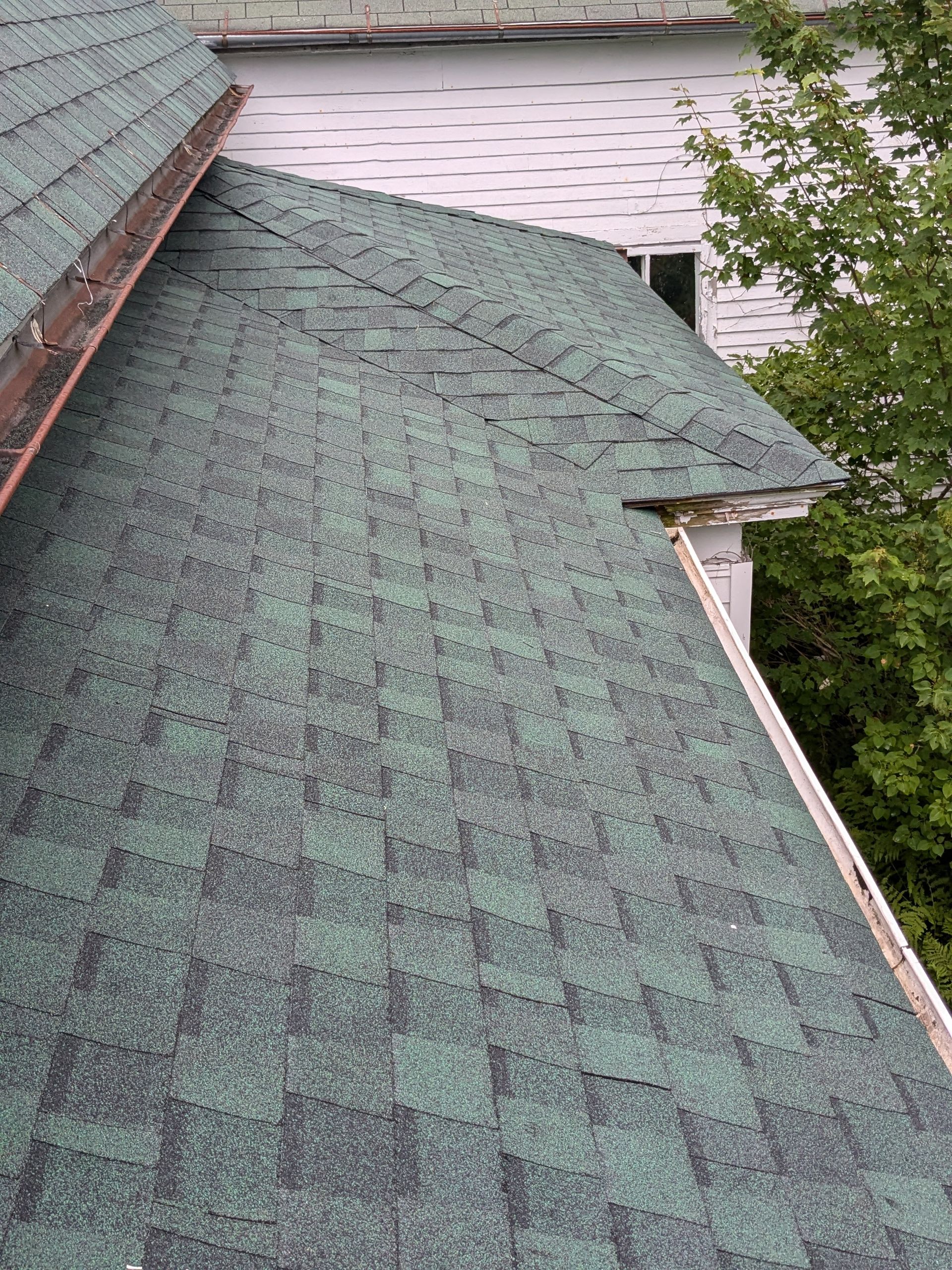 An elevated view of a green shingled roof featuring a valley and a copper gutter against a white house wall.