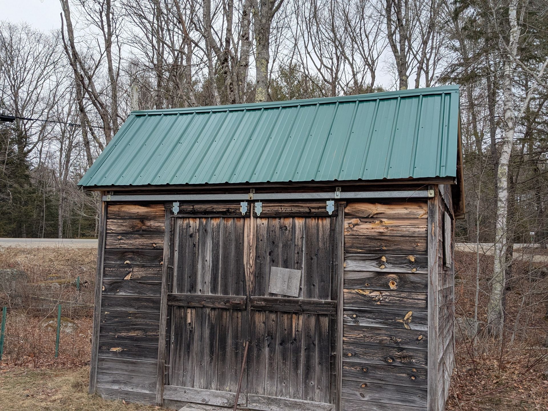 A small, weathered wooden shed with a green metal roof stands in a wooded area.