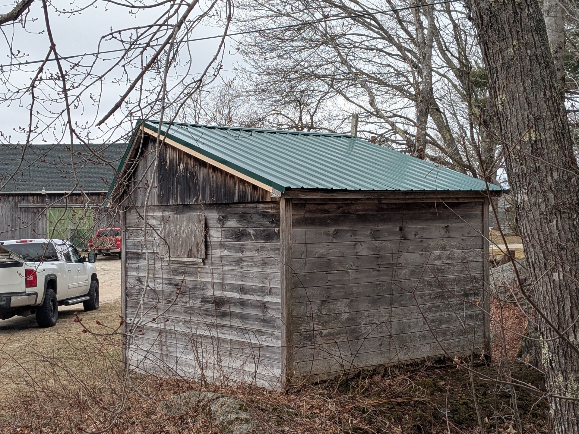 A small, weathered wooden shed with a new green metal roof sits in a yard with a white pickup truck in the background.