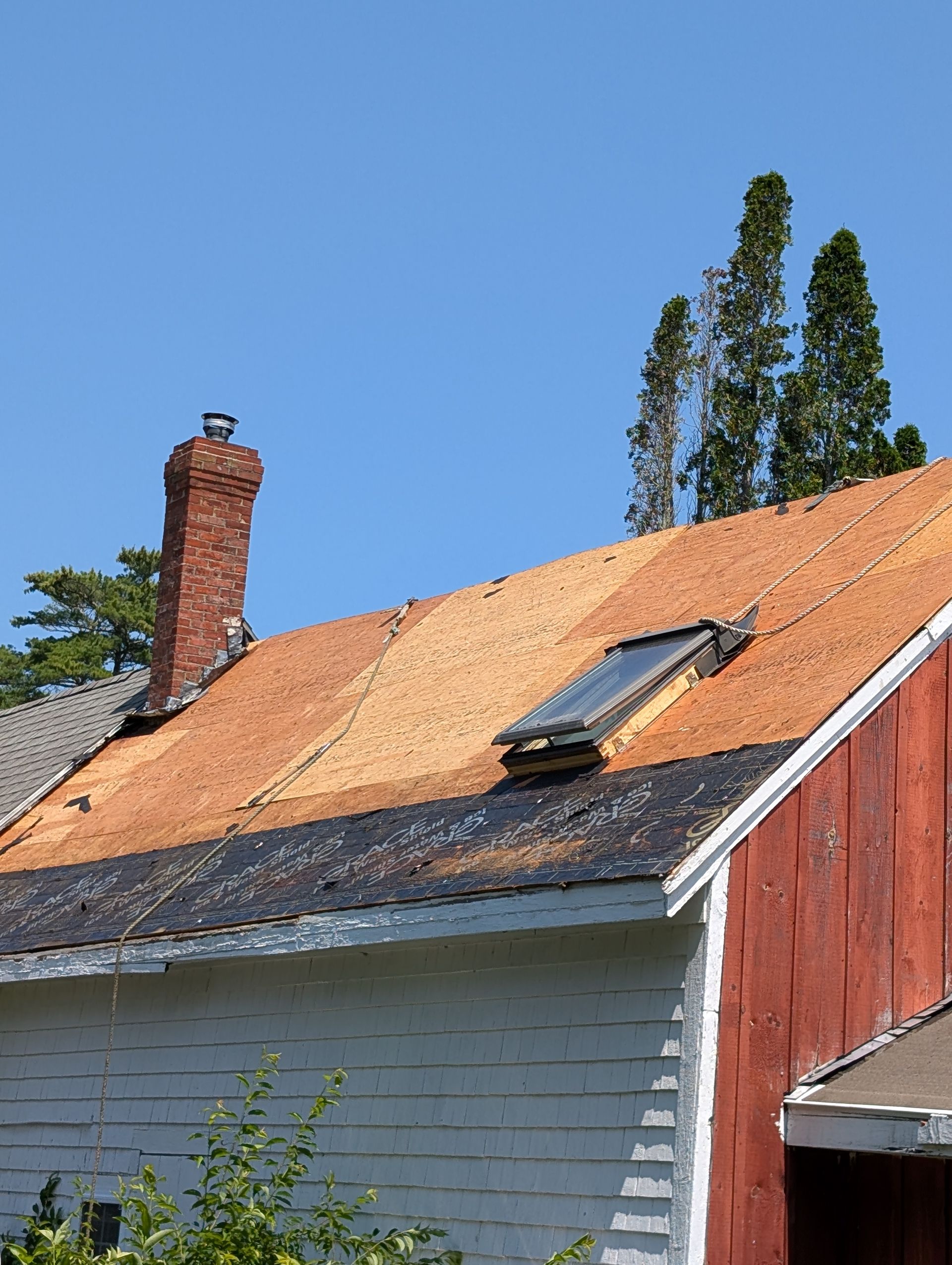 A section of a house roof undergoing repairs, showing bare plywood, a skylight, a brick chimney, and black roofing felt.