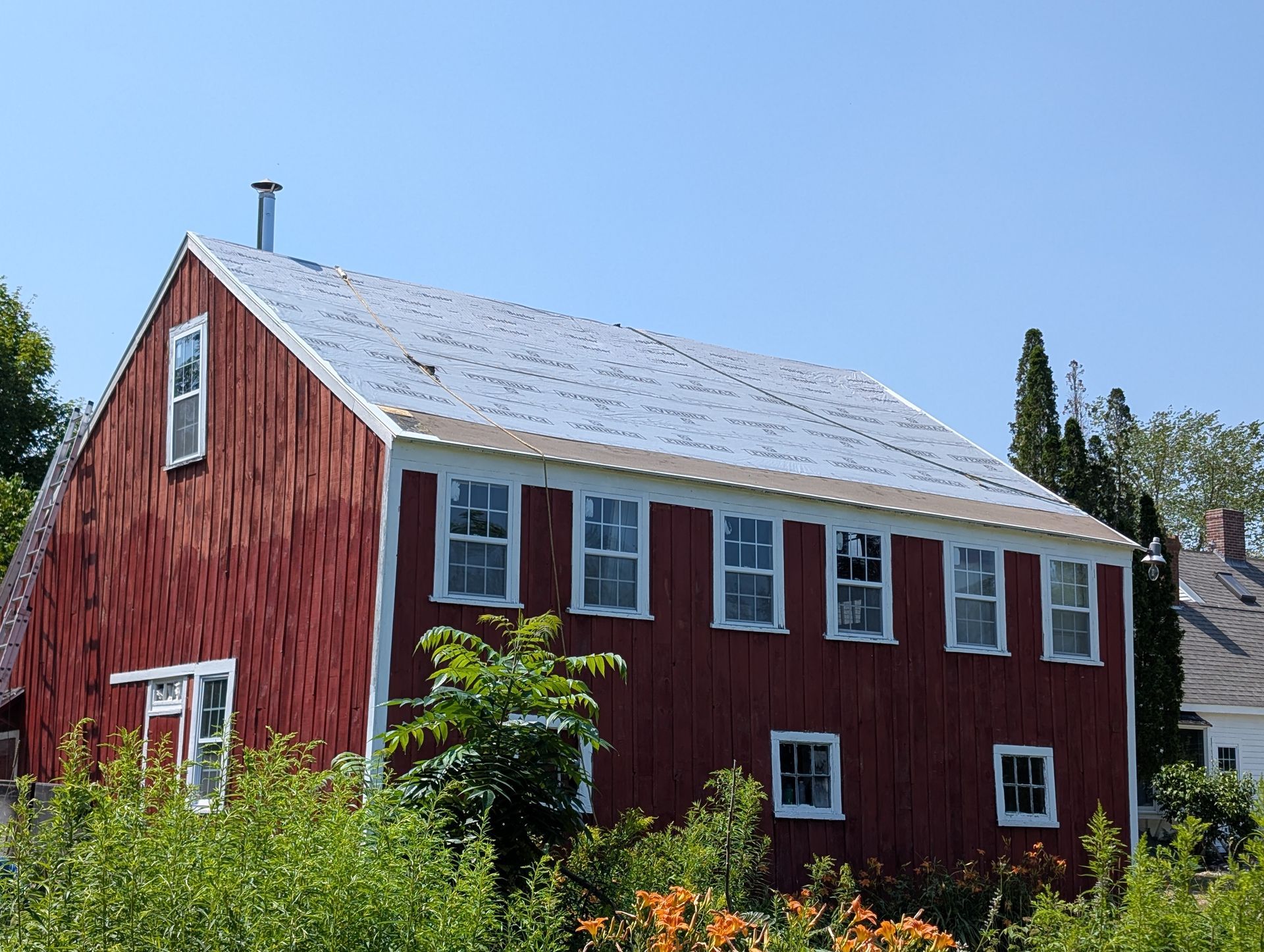 A two-story red barn with white trim and a light-colored roof surrounded by greenery under a clear blue sky.