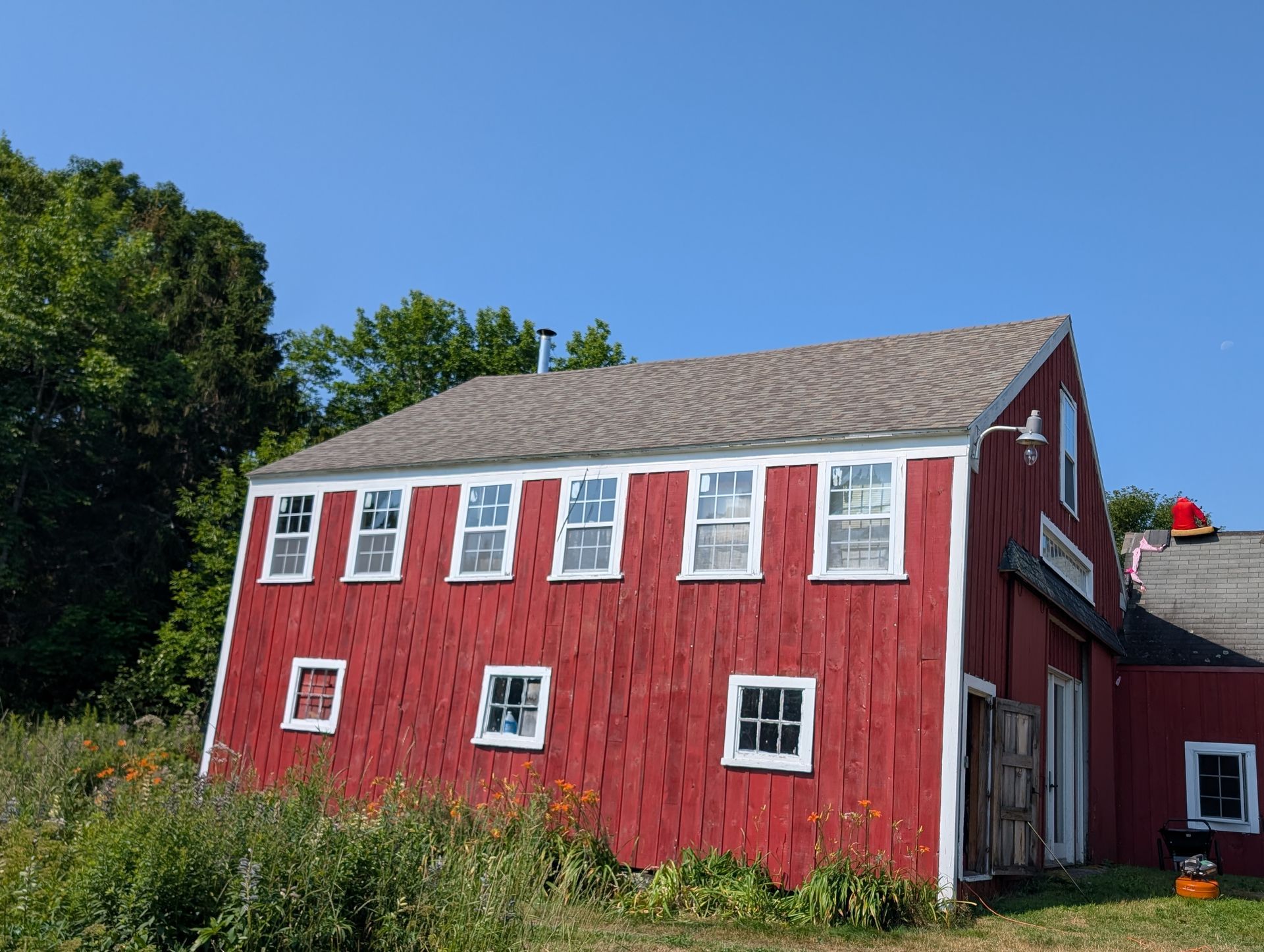 A red barn with white-trimmed windows and a gray shingled roof sits in a field under a clear blue sky.